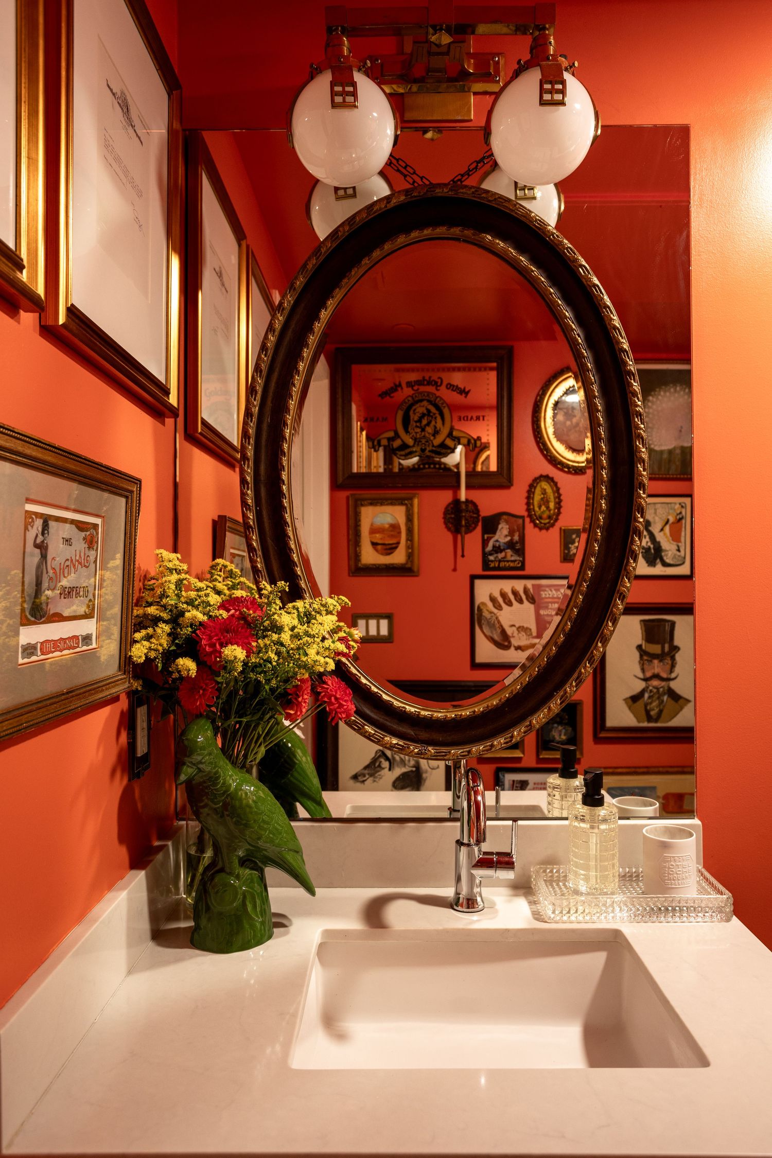 Vibrant red bathroom with oval mirror, white sink, floral arrangement, and eclectic framed art gallery wall.
