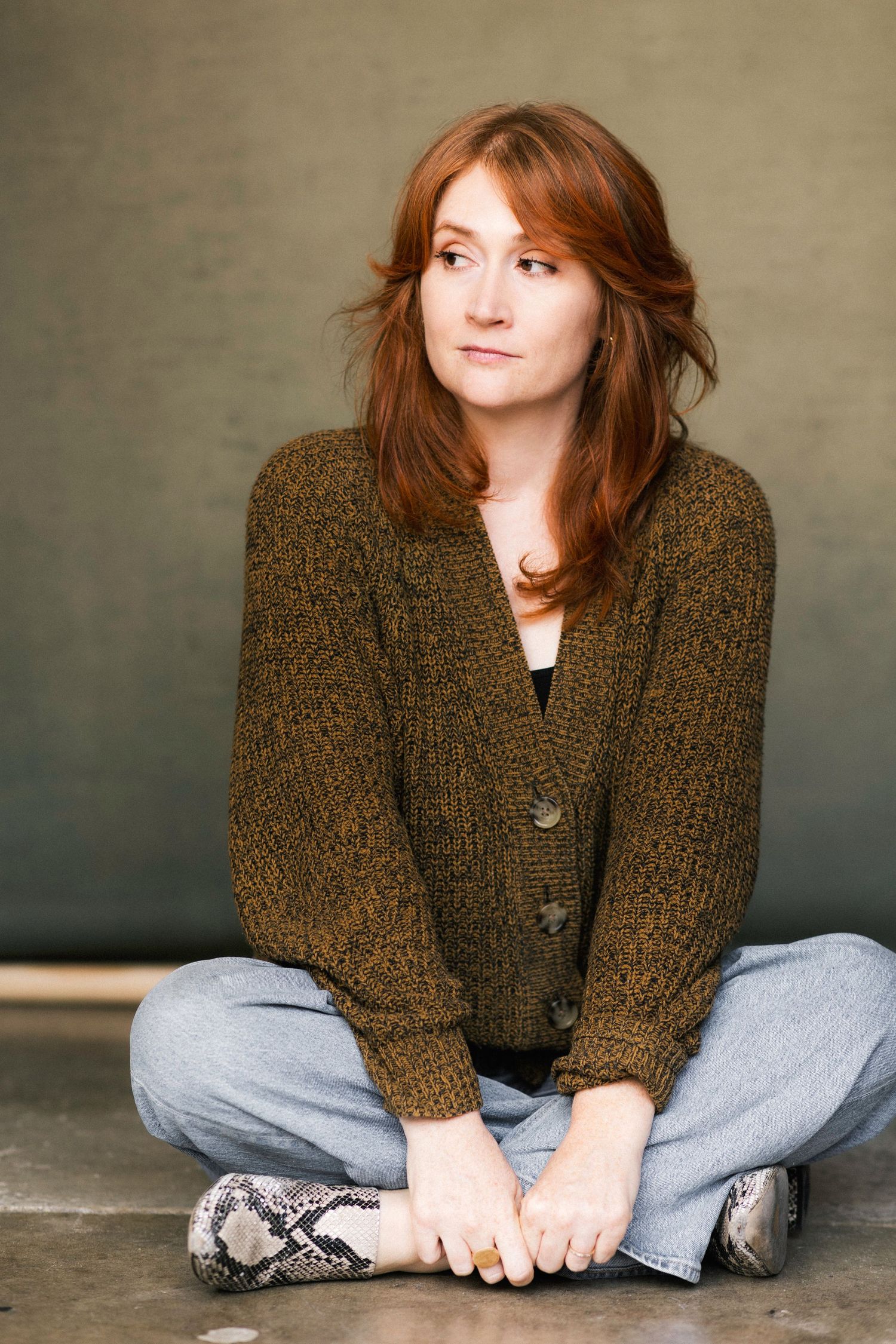 Young woman with red hair sitting cross-legged on floor wearing olive cardigan and jeans with snake print shoes.