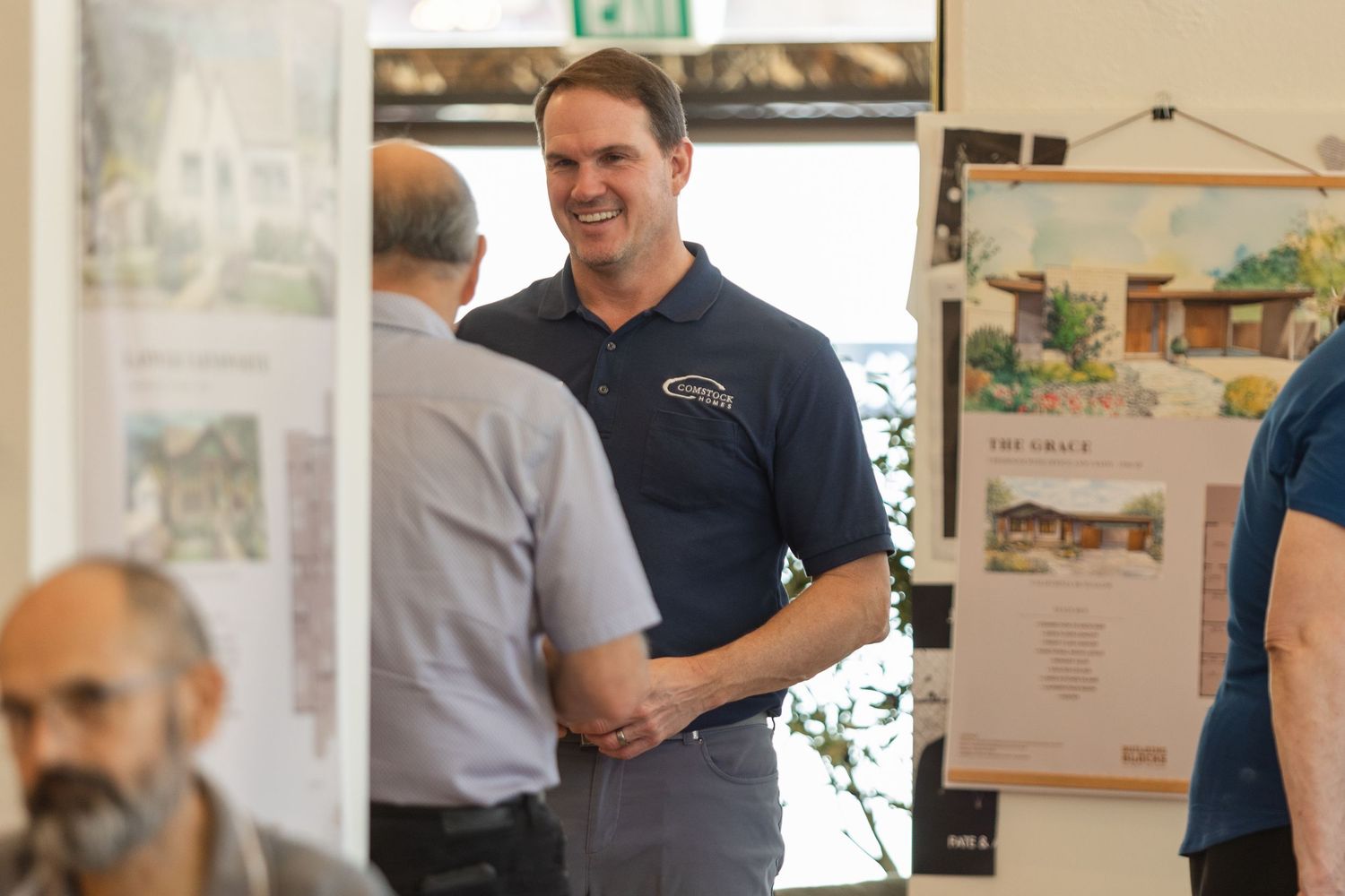 Two people having a business conversation in an office space with architectural drawings displayed on the wall.