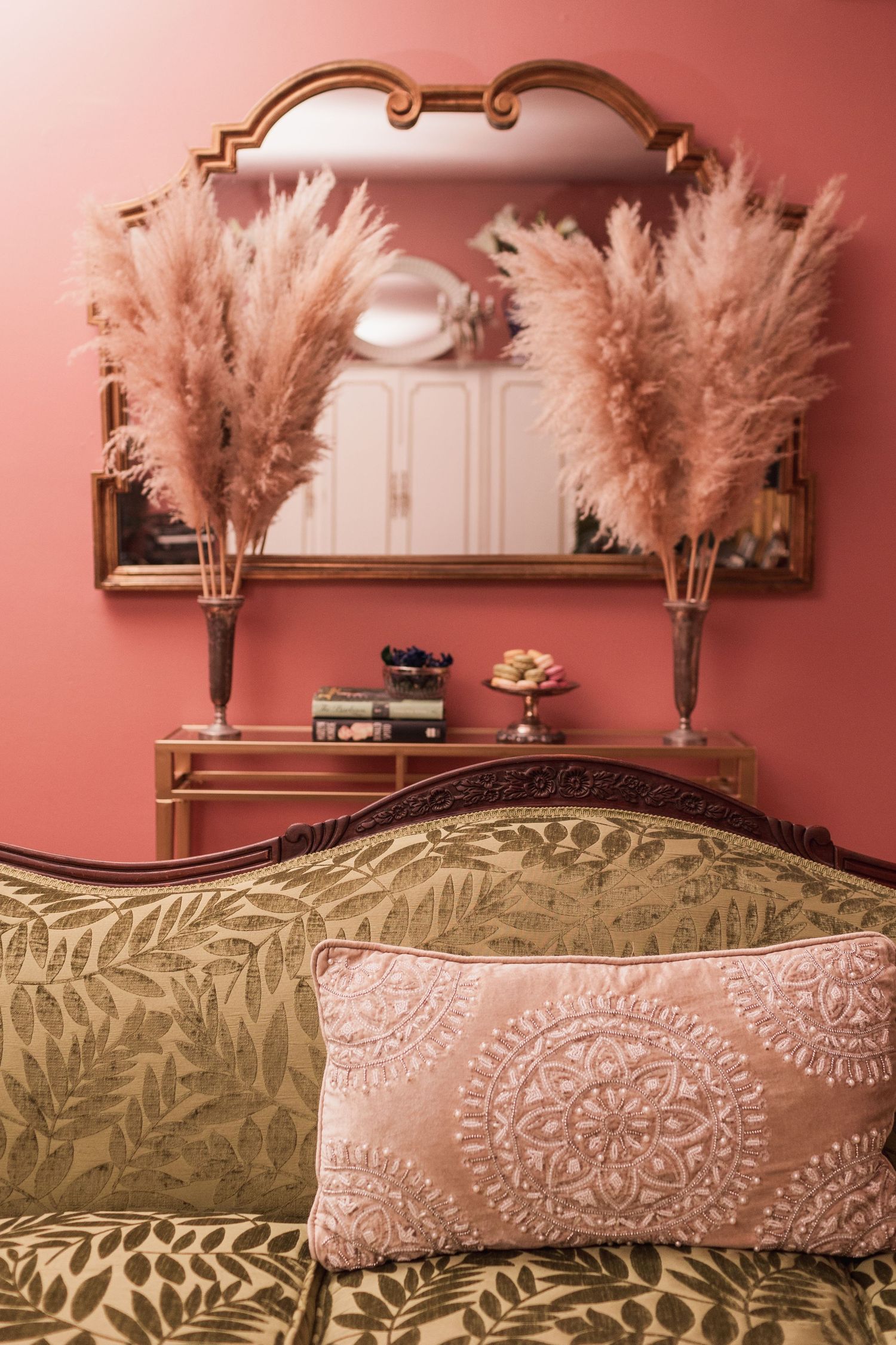 Elegant bedroom with pink walls, gold mirror, pampas grass decor, and green floral sofa with pink embroidered pillow.