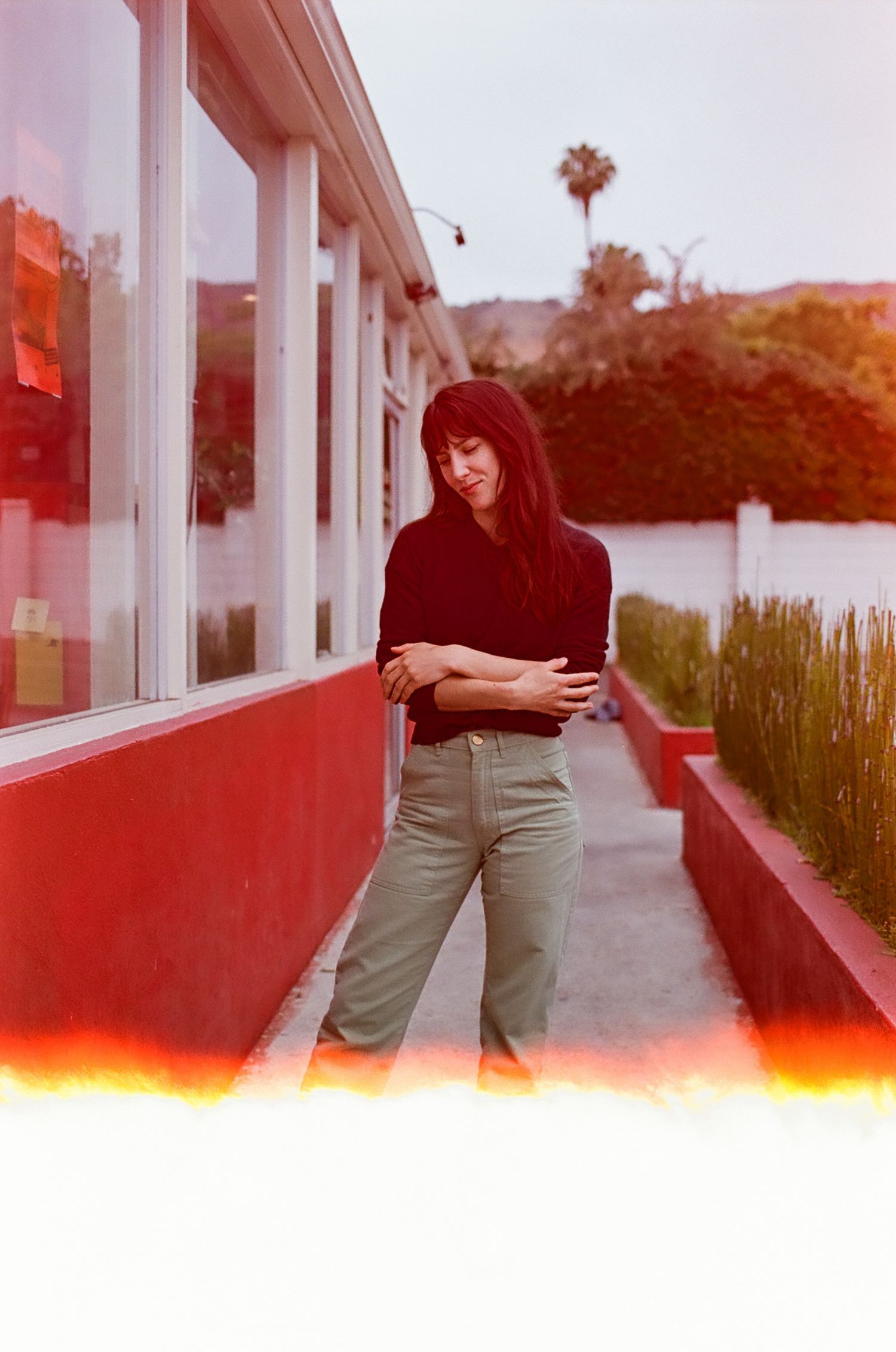 A young woman with dark hair in a black sweater and gray pants stands outside a red building with large windows.