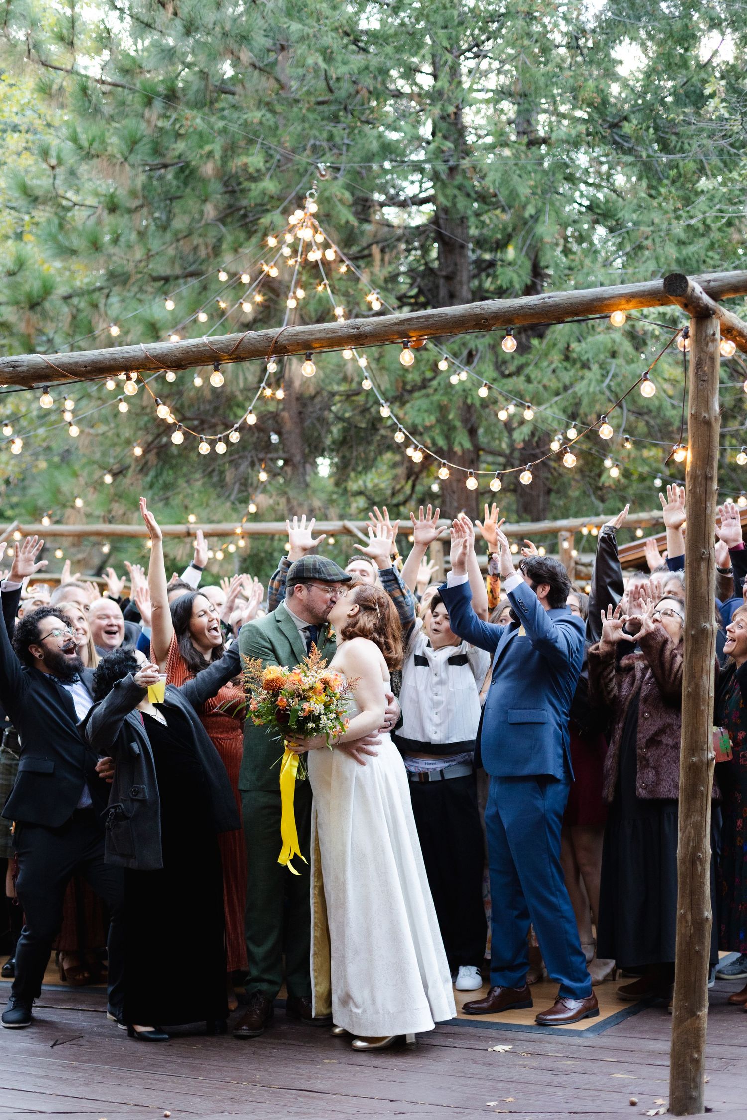 Large wedding celebration with guests raising hands under wooden pergola outdoors.