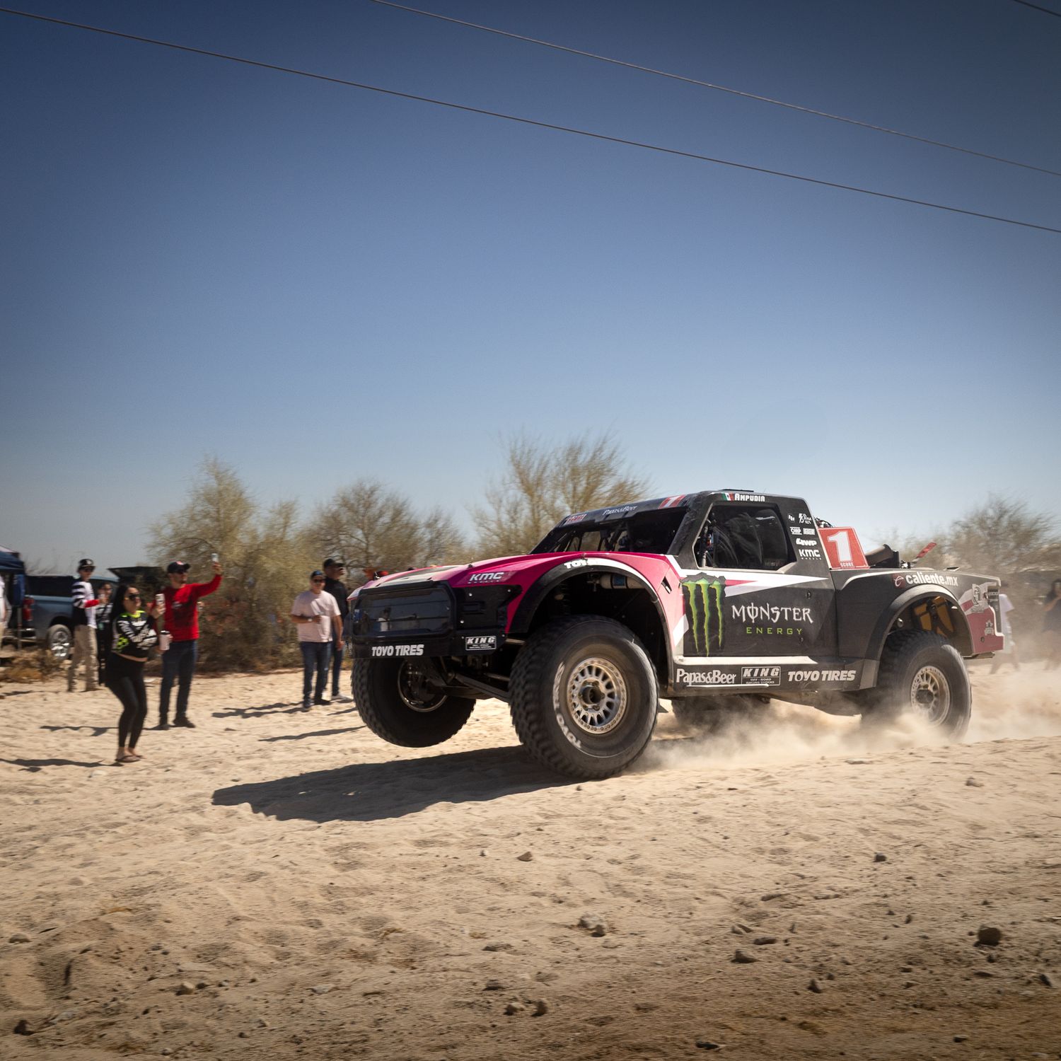 “Monster Energy Trophy Truck racing past spectators on a dusty San Felipe 250 desert straightaway.”