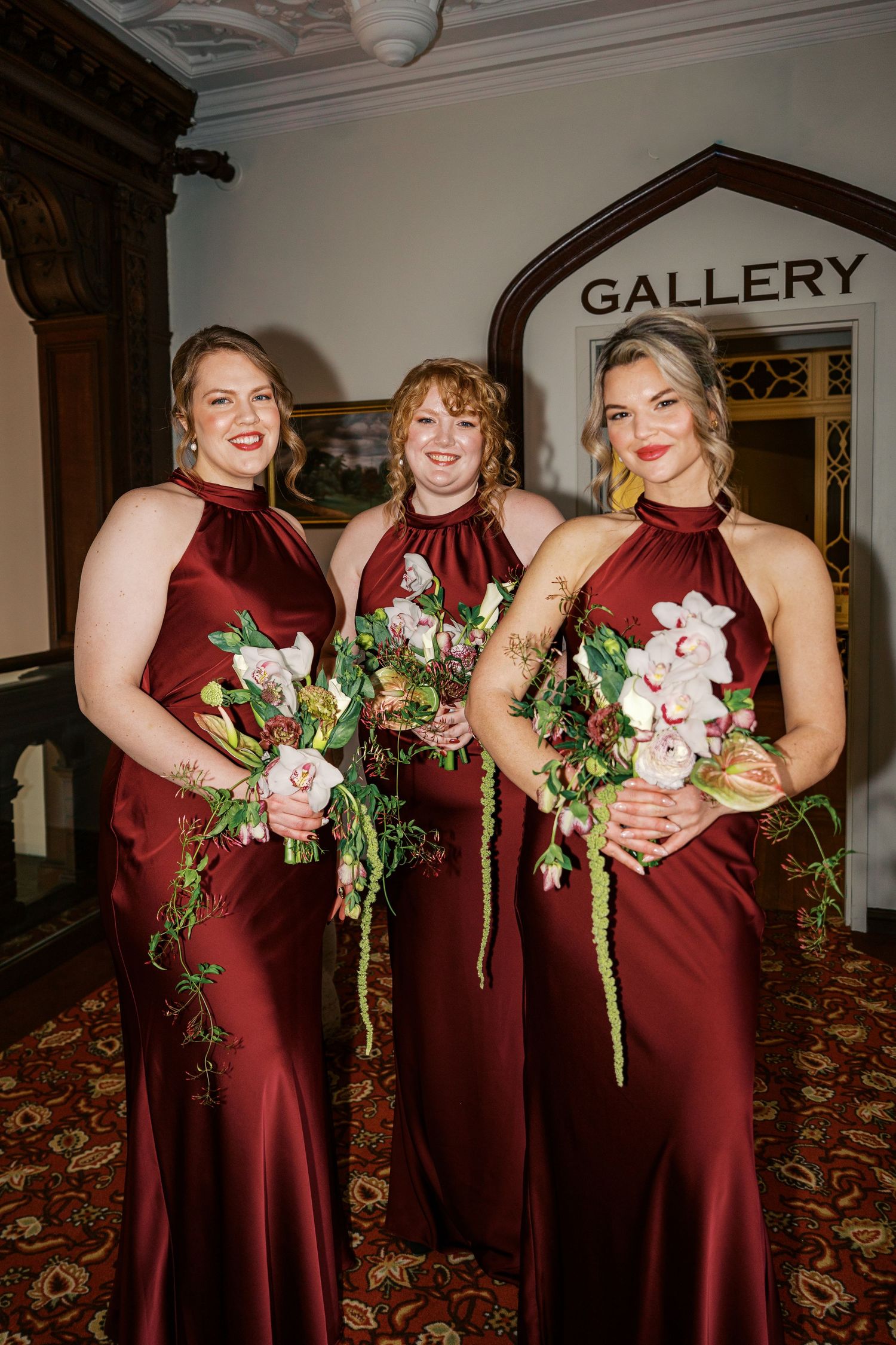 Bridesmaids in burgundy dresses pose together holding floral bouquets in an elegant indoor gallery setting.