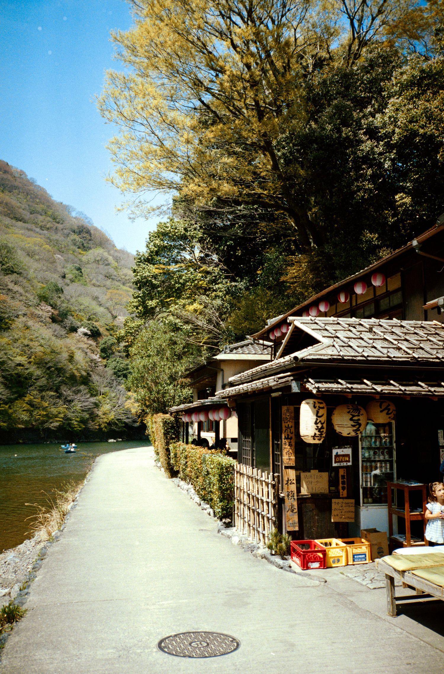 Traditional Japanese buildings line a riverside path surrounded by autumn trees.