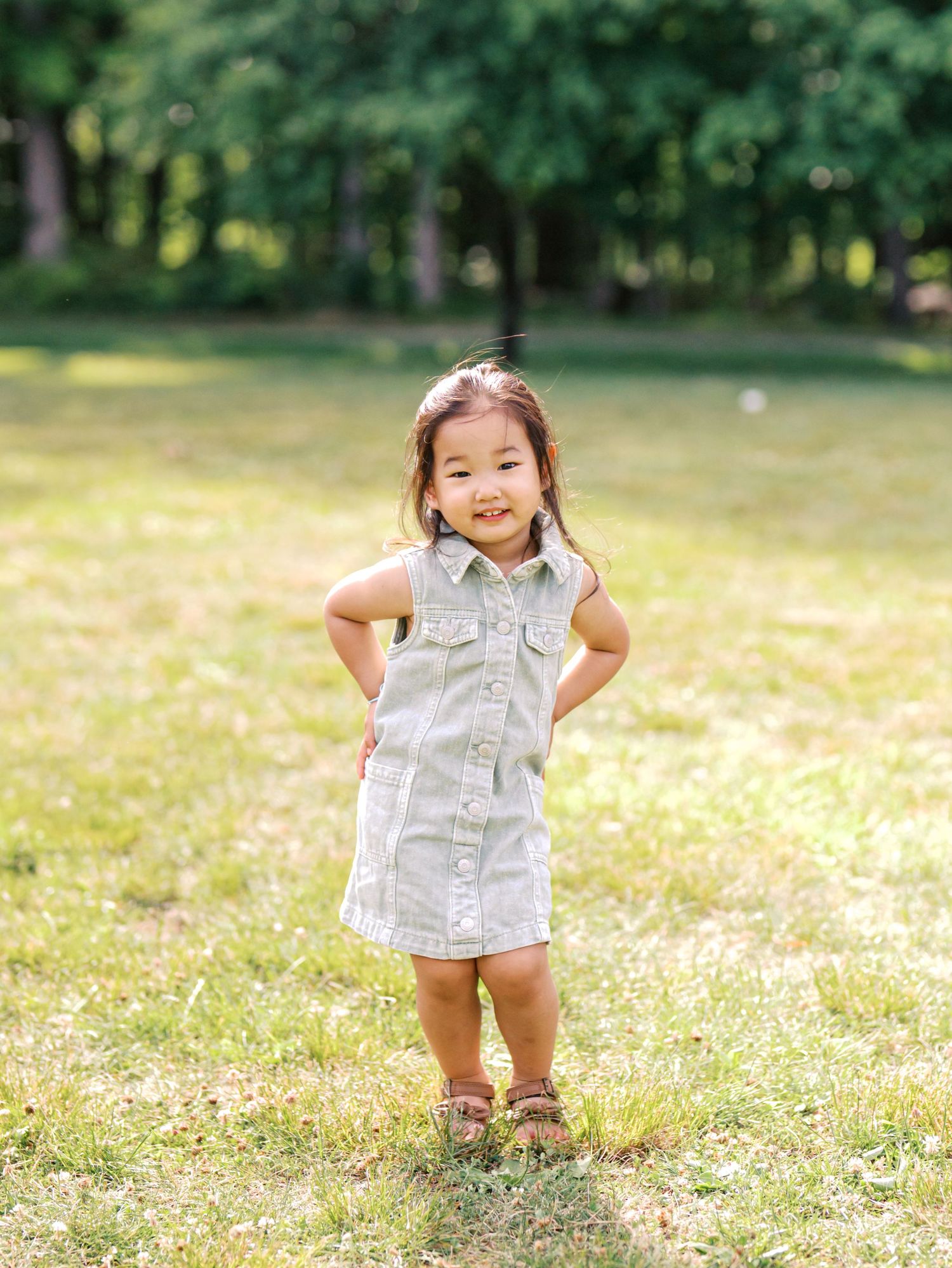 A smiling toddler girl in a sage green dress stands confidently in a sunny park with hands on hips.