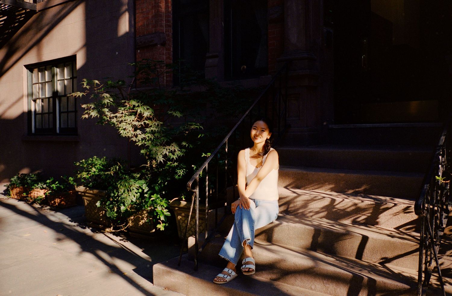 Person sits on sunlit brownstone steps in casual denim, surrounded by greenery and shadows on a city street.