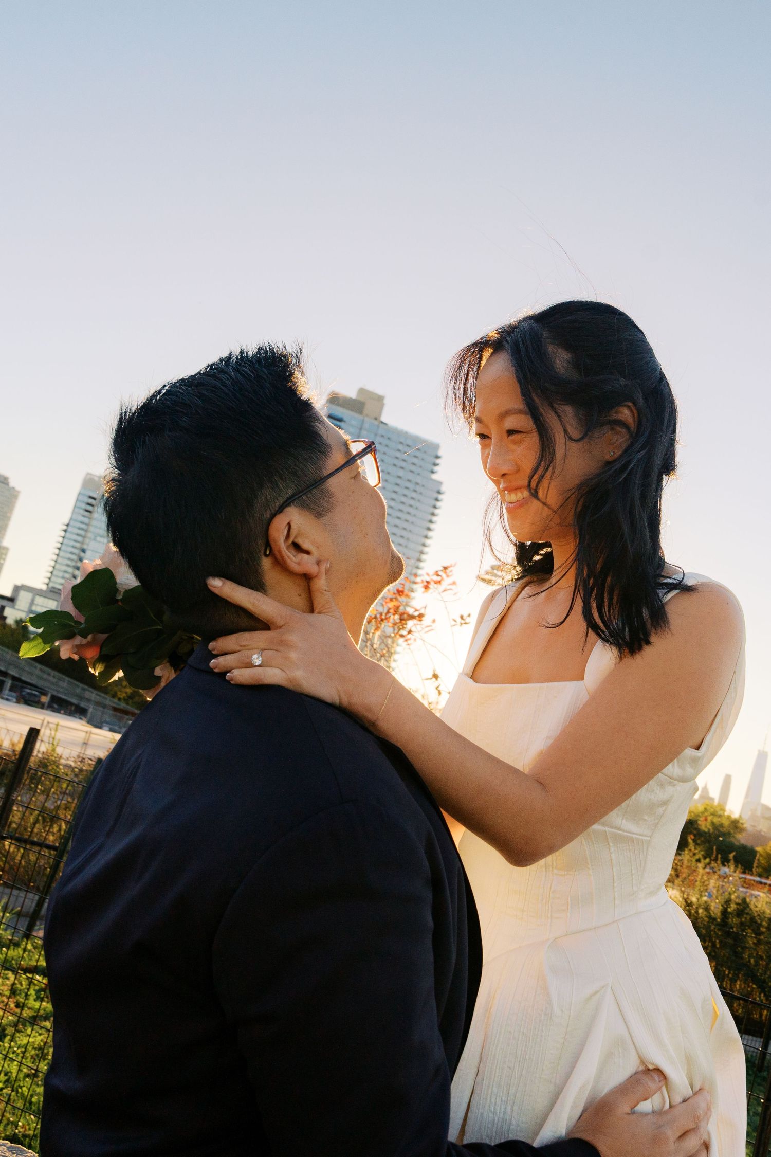 A romantic sunset engagement photo with a couple embracing outdoors against a city skyline backdrop.