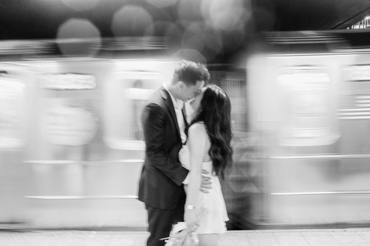 Black and white photo of a couple kissing in wedding attire with a blurred train passing behind them.