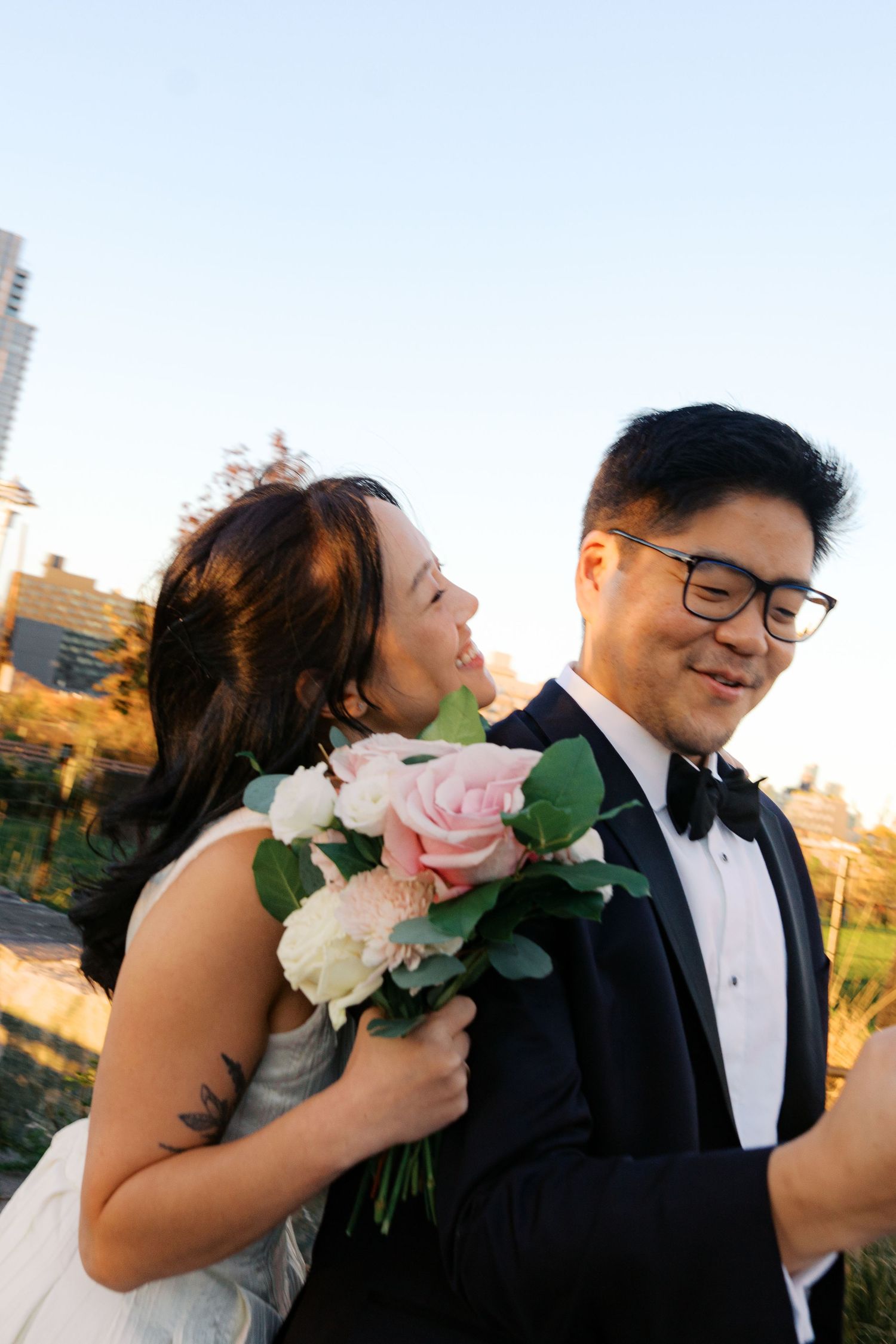 A couple shares romantic moments in formal wedding attire while holding a pink and white rose bouquet at sunset.