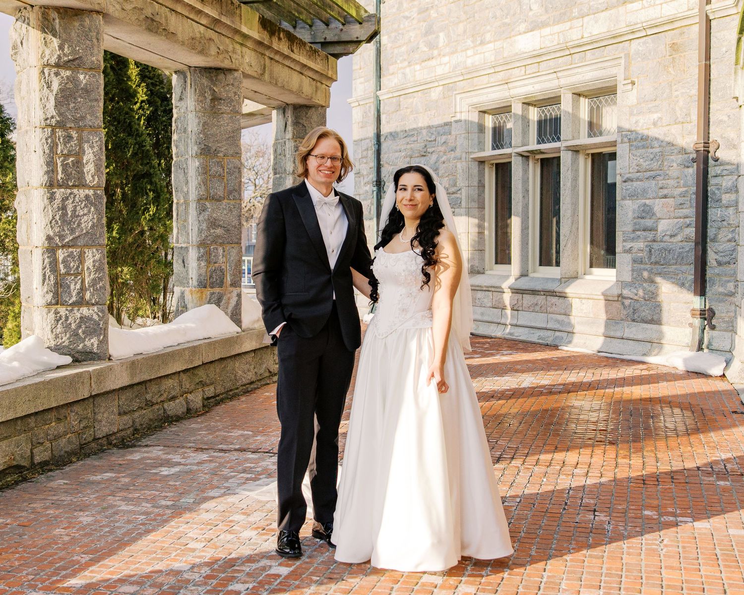 A wedding couple walks along a stone building exterior with elegant architectural details and brick pathways.