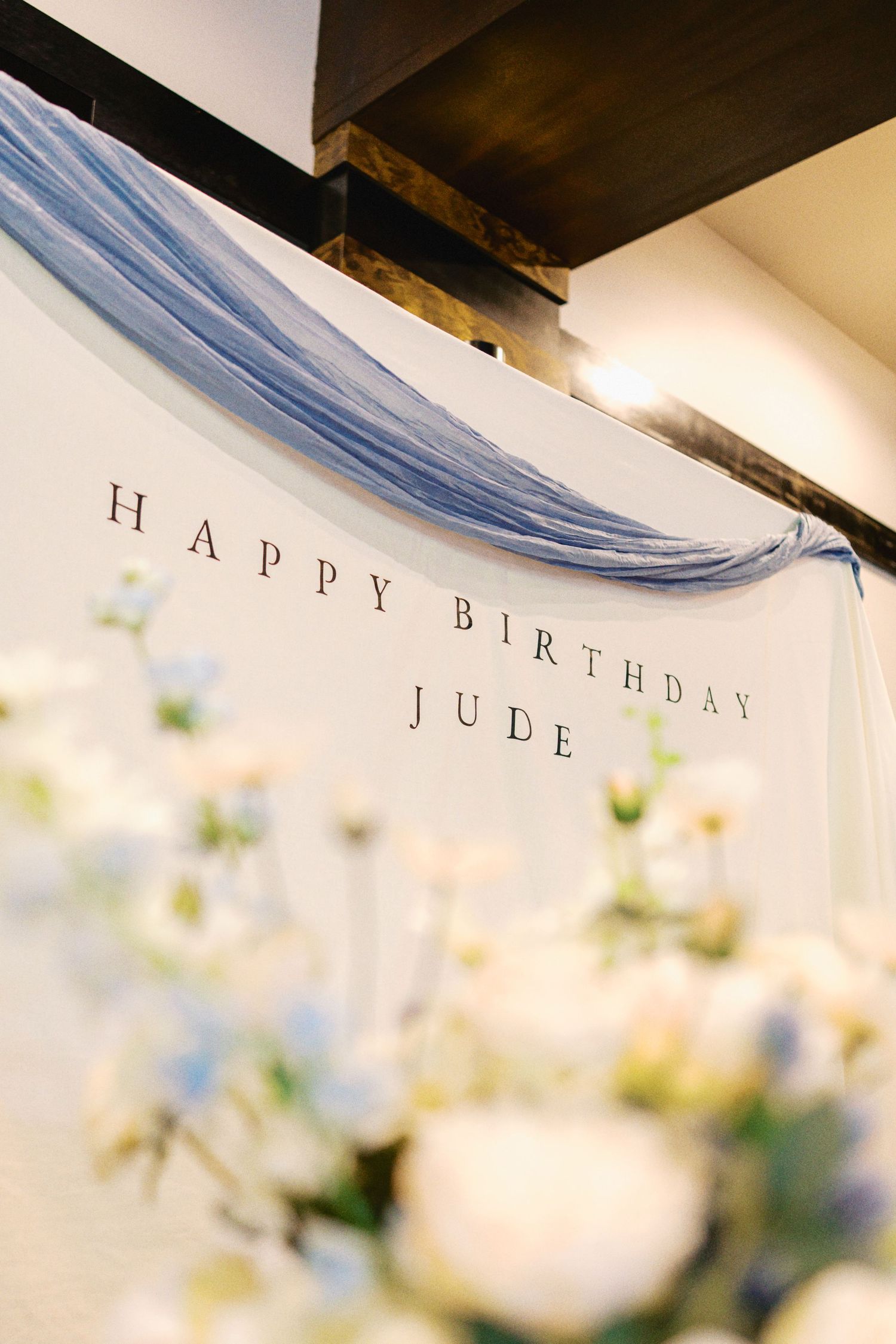 Happy Birthday Jude banner with blue draping and white floral arrangement at a birthday celebration.