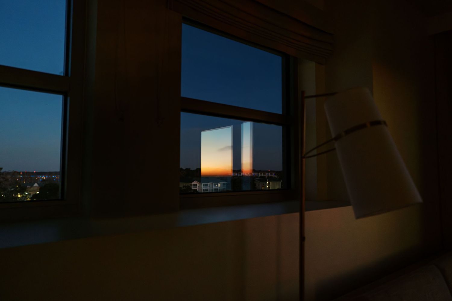 A floor lamp illuminates a dark room with a window view of city lights and sunset in the distance.