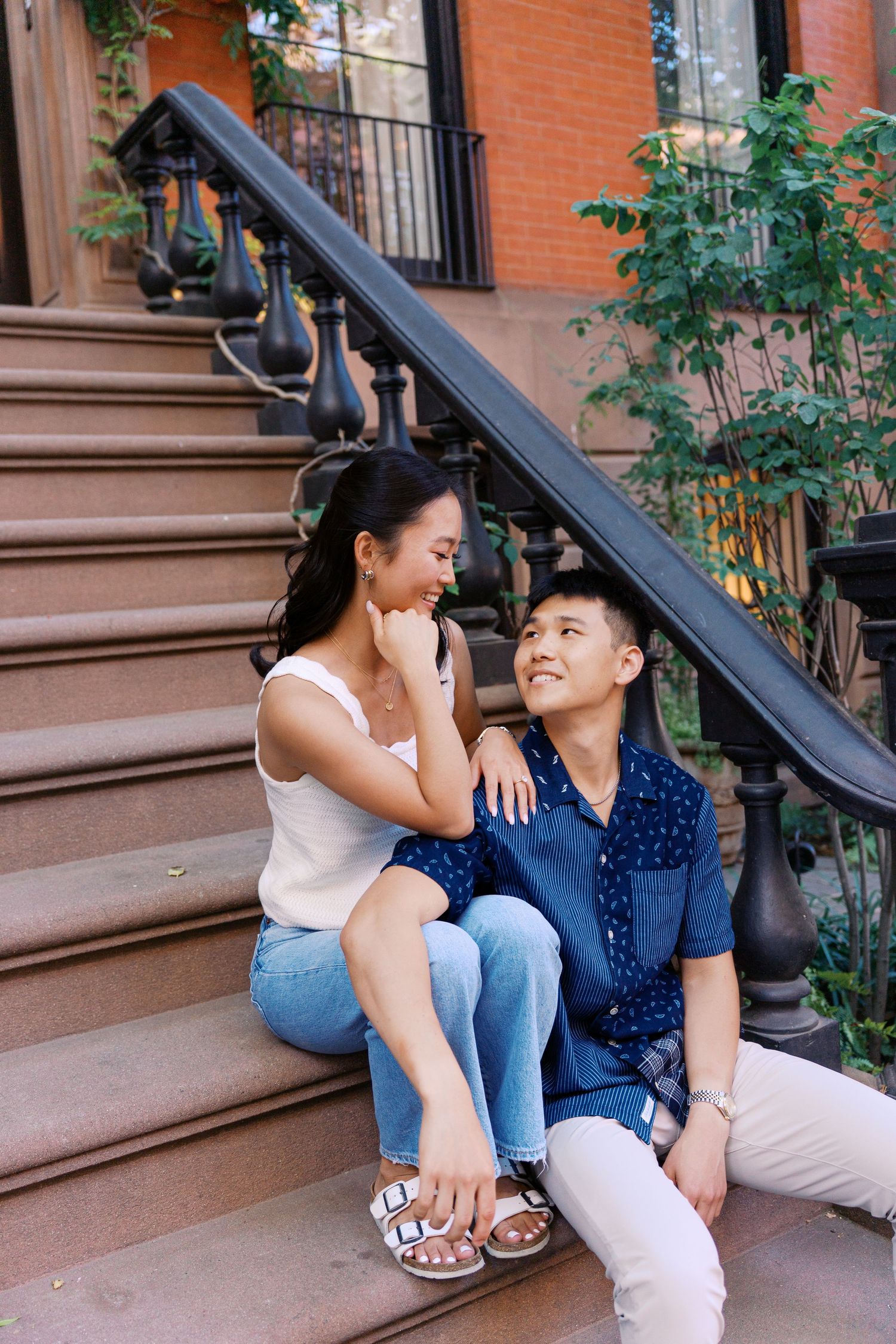 Two people share a joyful moment while sitting on brownstone steps in casual summer outfits.