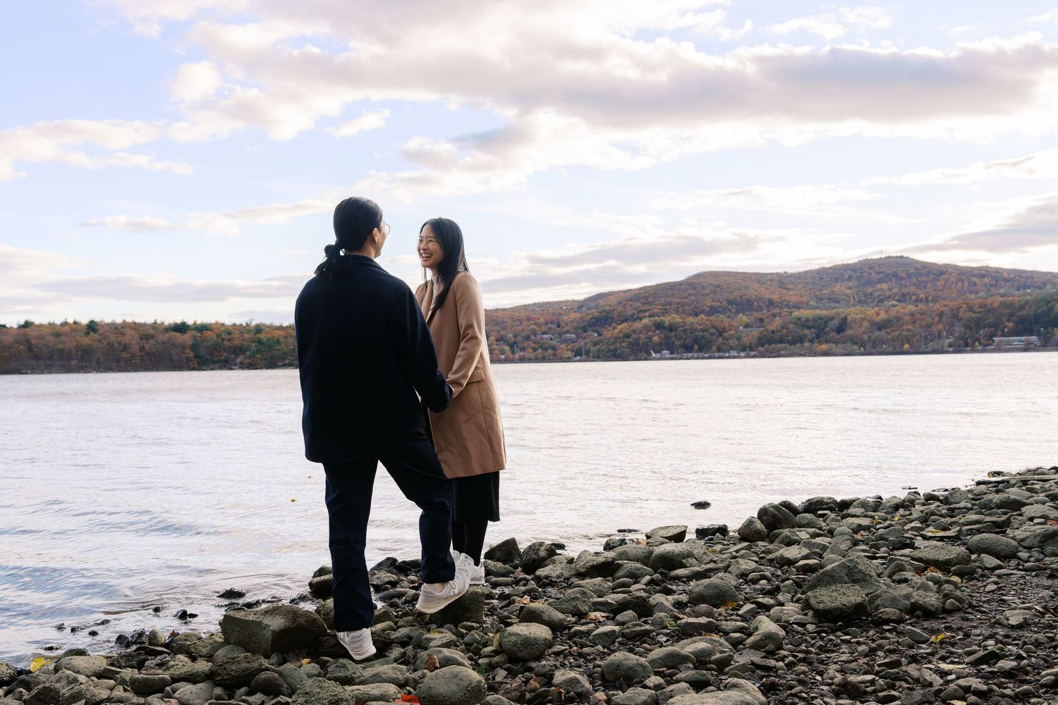 Two people sit together on rocky shoreline overlooking a serene lake with mountains in the background during sunset.