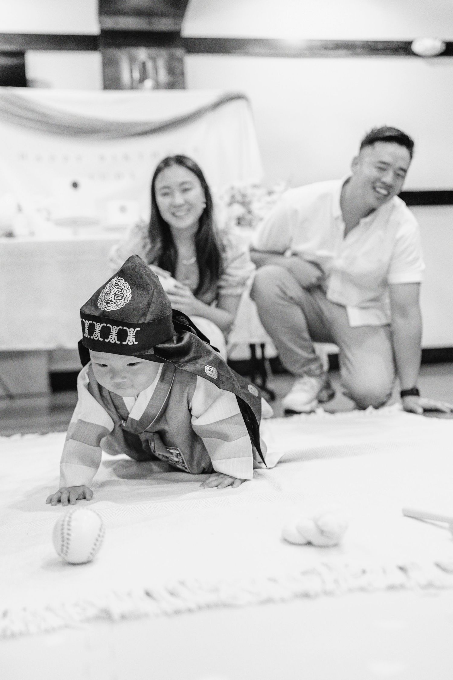 Baby in traditional Korean hanbok crawling during doljabi ceremony while parents smile joyfully.