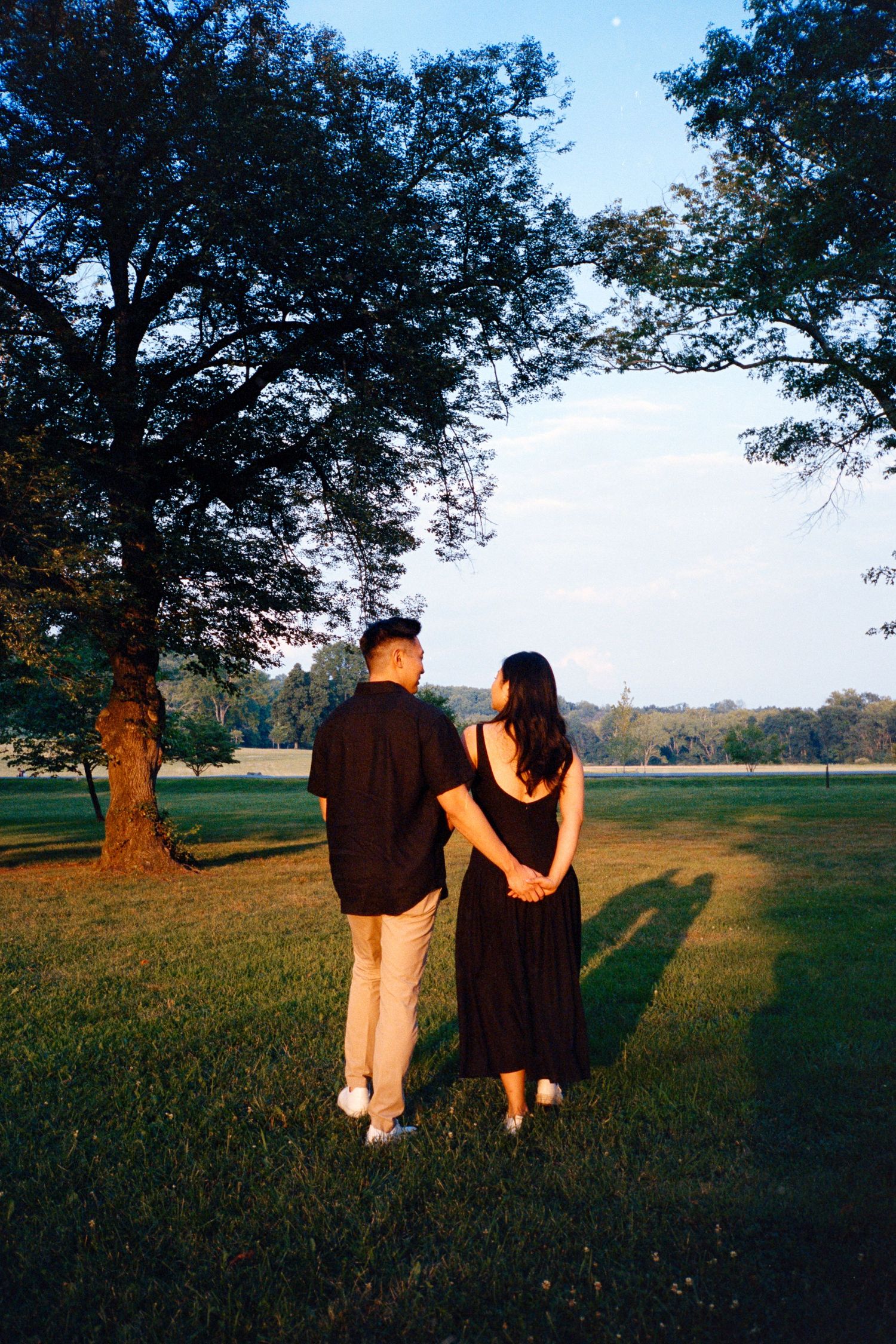Two figures walking together across a grassy field at sunset with trees silhouetted in the background.