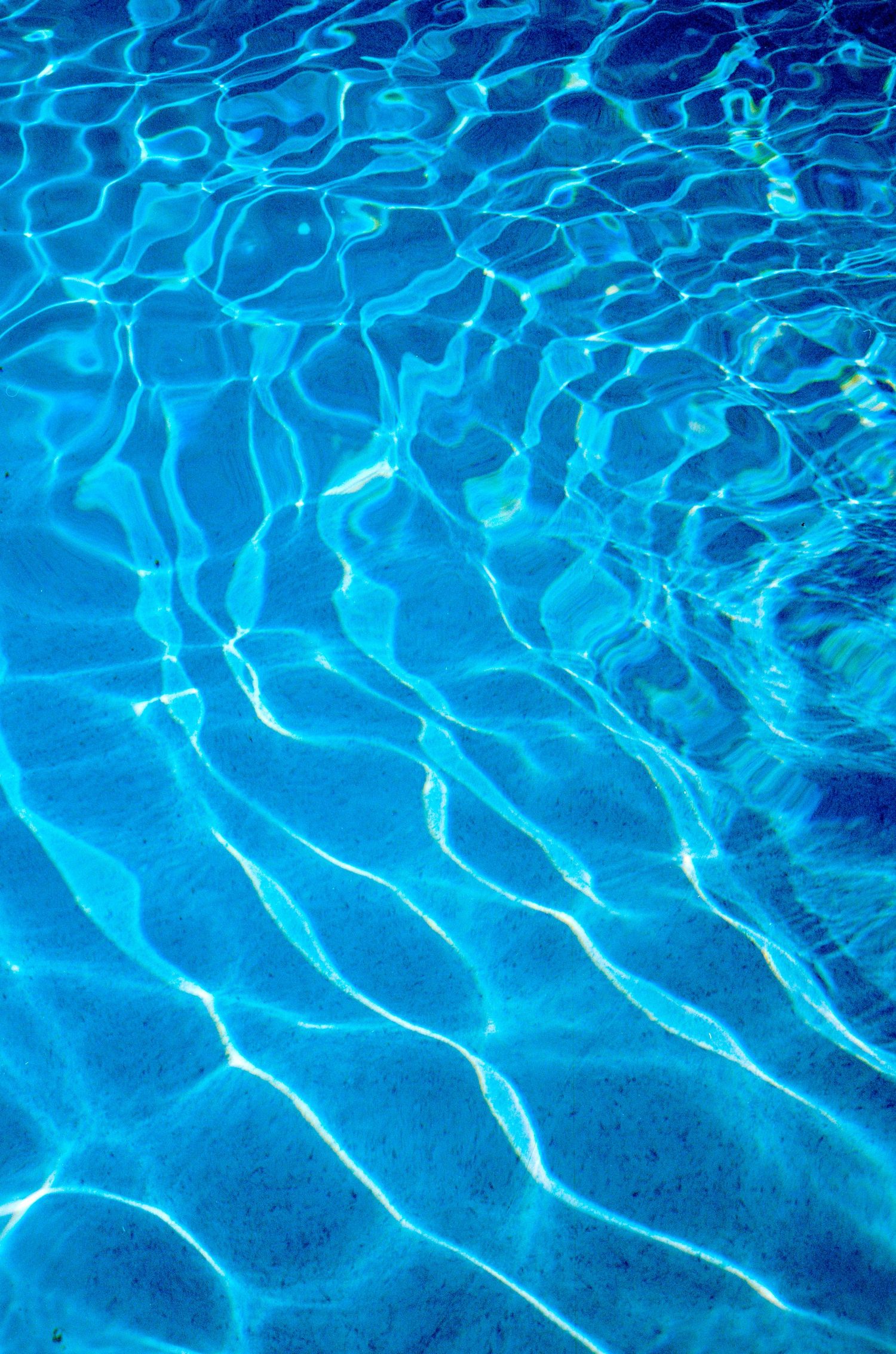 Rippling blue water surface with sunlight creating bright reflections and wave patterns in a swimming pool.