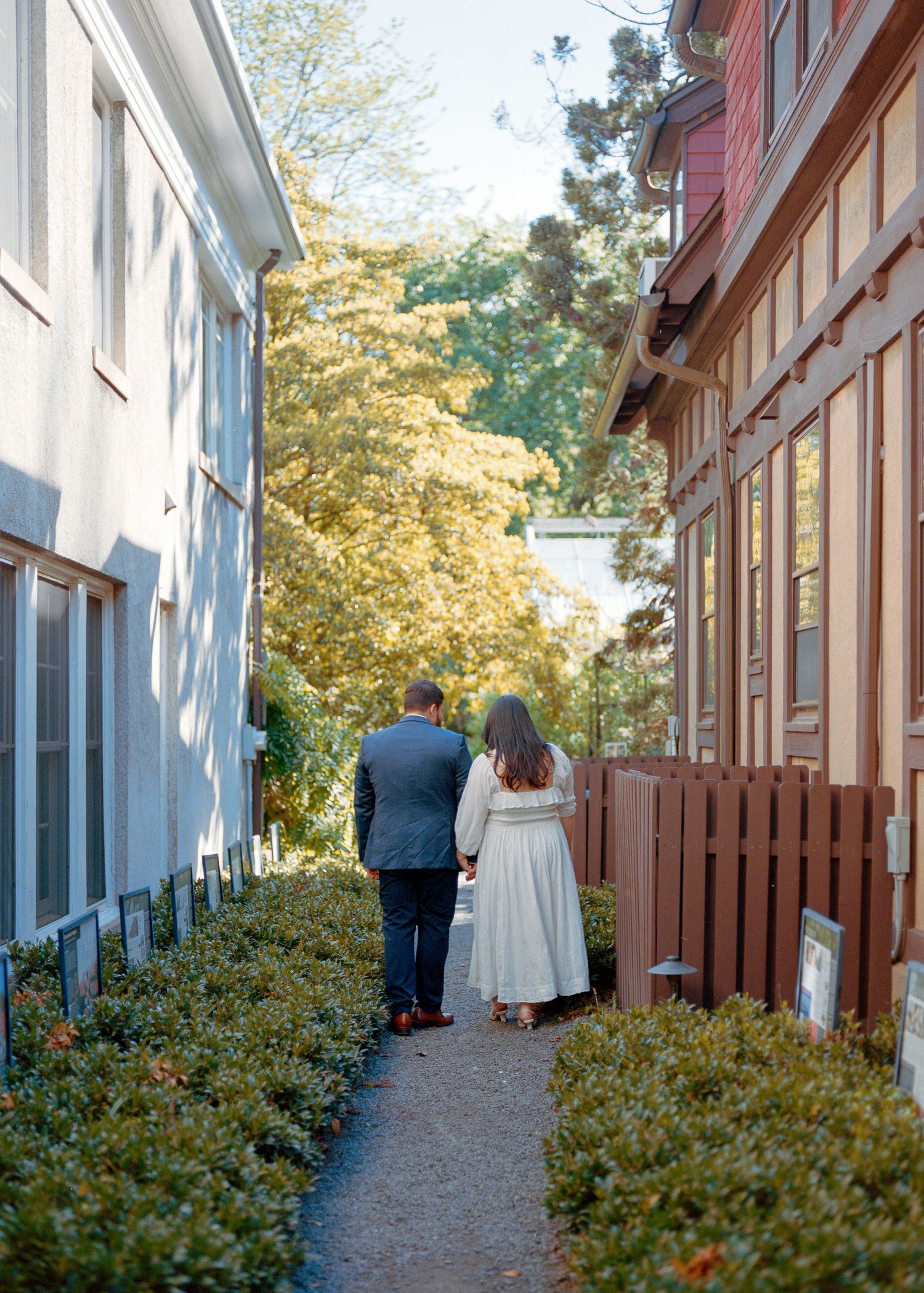 A narrow pathway between historic buildings with shrubs and autumn foliage.