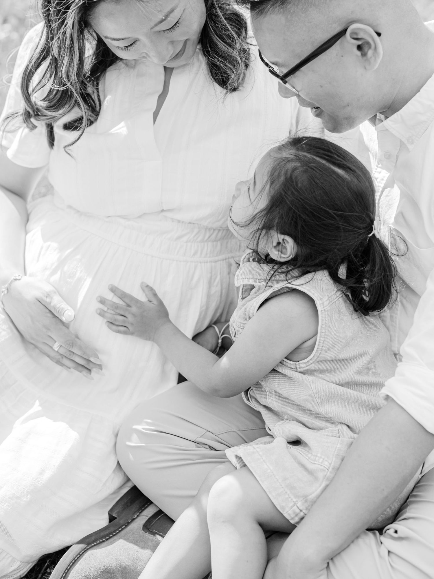 Black and white maternity photo of pregnant woman with husband and toddler daughter touching her belly.