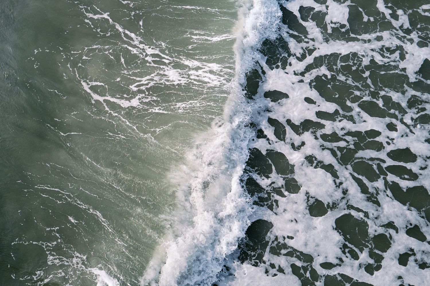 Aerial view of ocean waves crashing against dark rocks creating white foam patterns along the coastline.