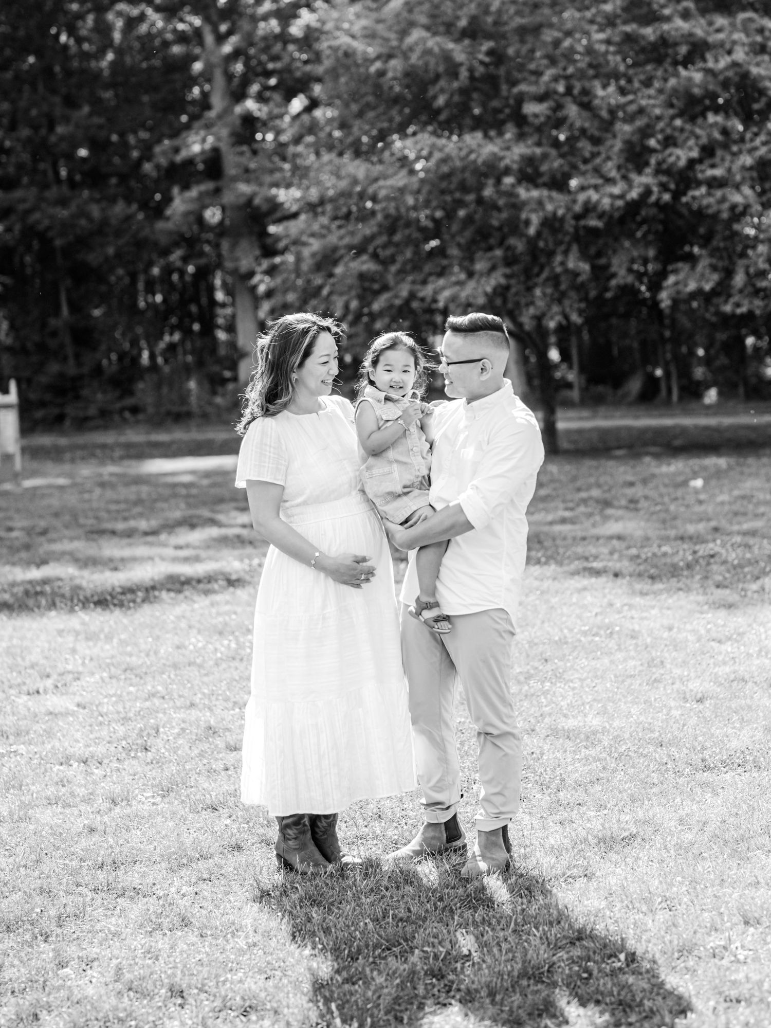 Black and white maternity photo of a pregnant woman in a white dress with her partner holding a toddler outdoors.