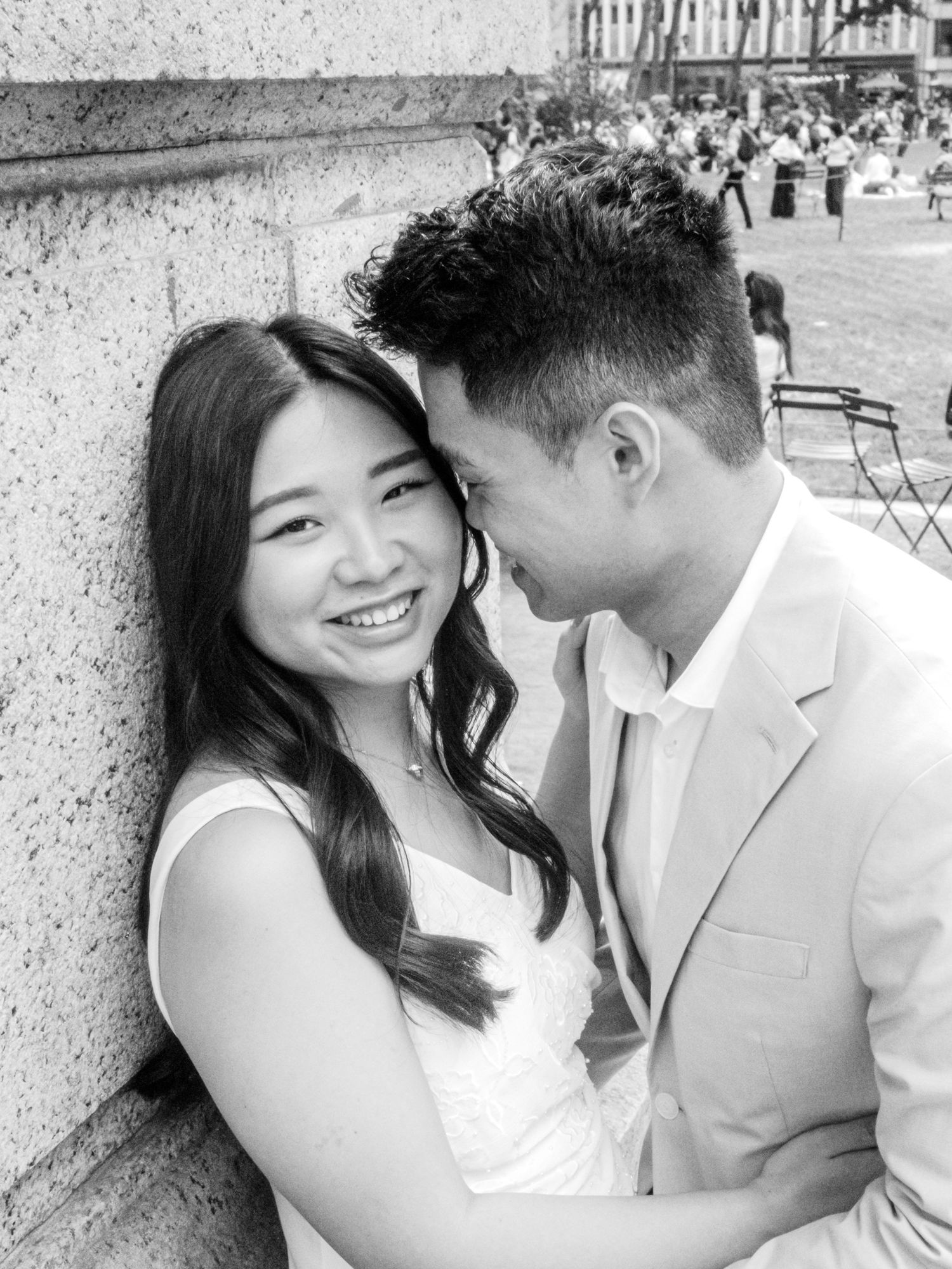 Black and white photo of a smiling couple in wedding attire leaning against a stone wall outdoors.