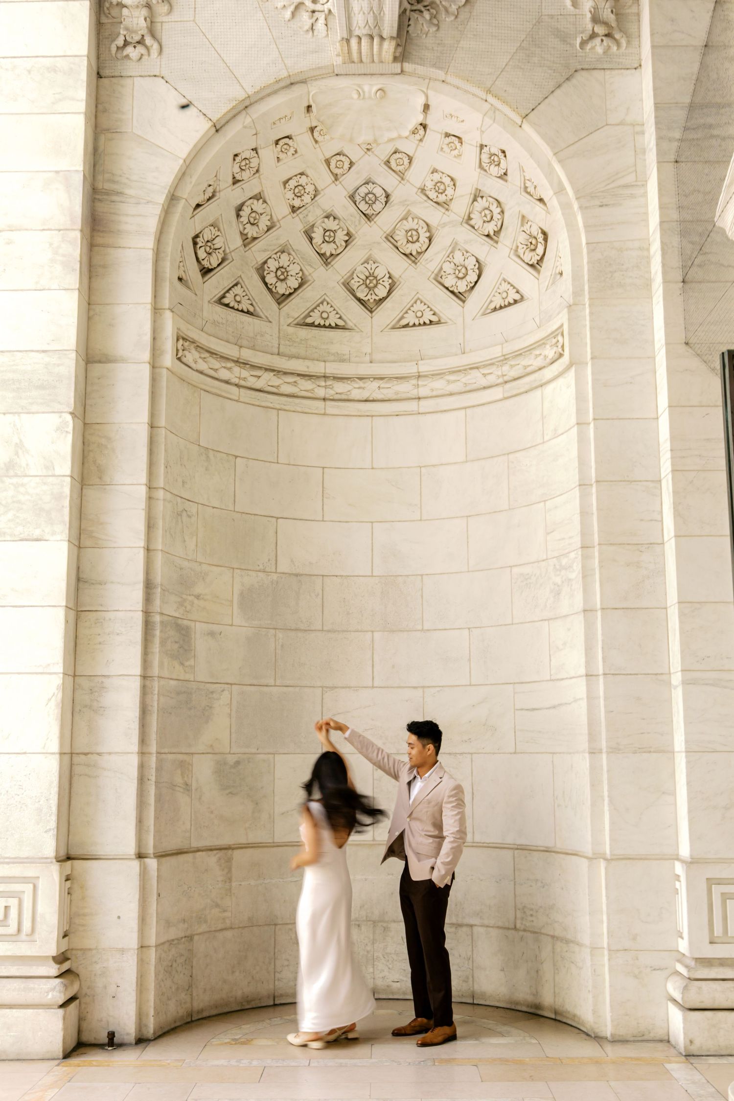 Couple dancing elegantly in front of ornate marble archway, woman in white gown being twirled by man in light suit.