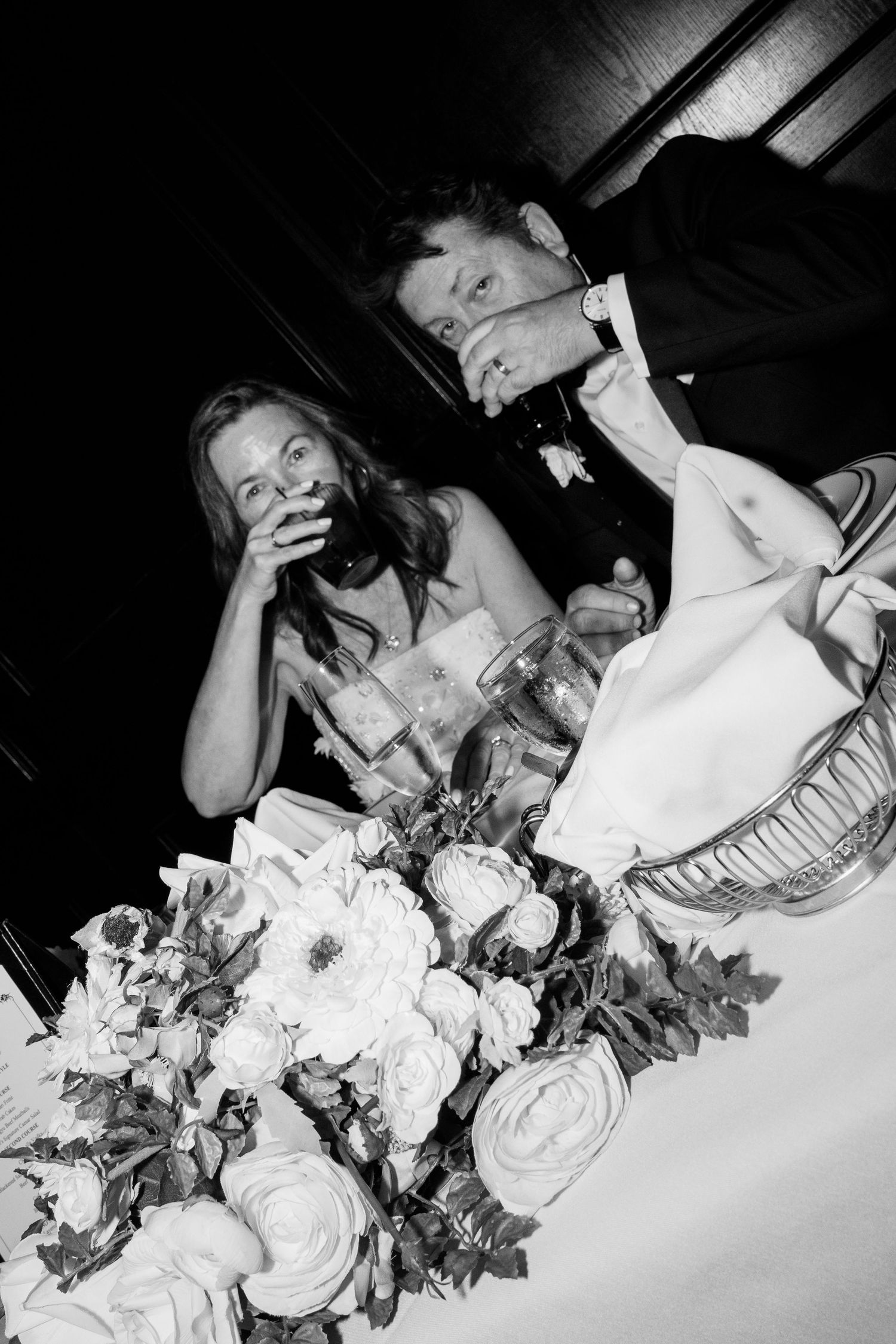Black and white photo of guests enjoying a wedding reception dinner with floral centerpieces.