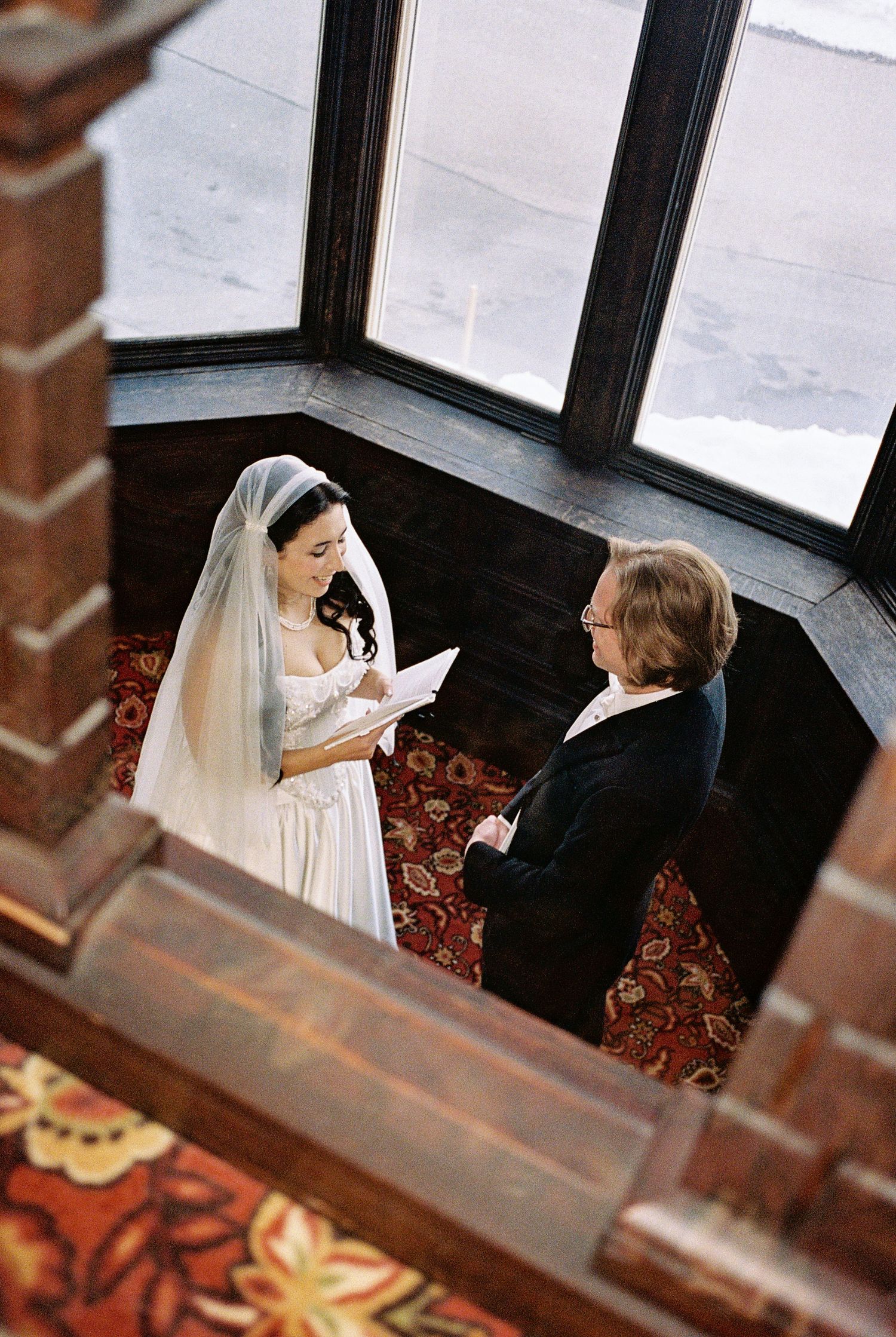 An overhead view captures a wedding ceremony moment on a patterned red carpet beside large windows.