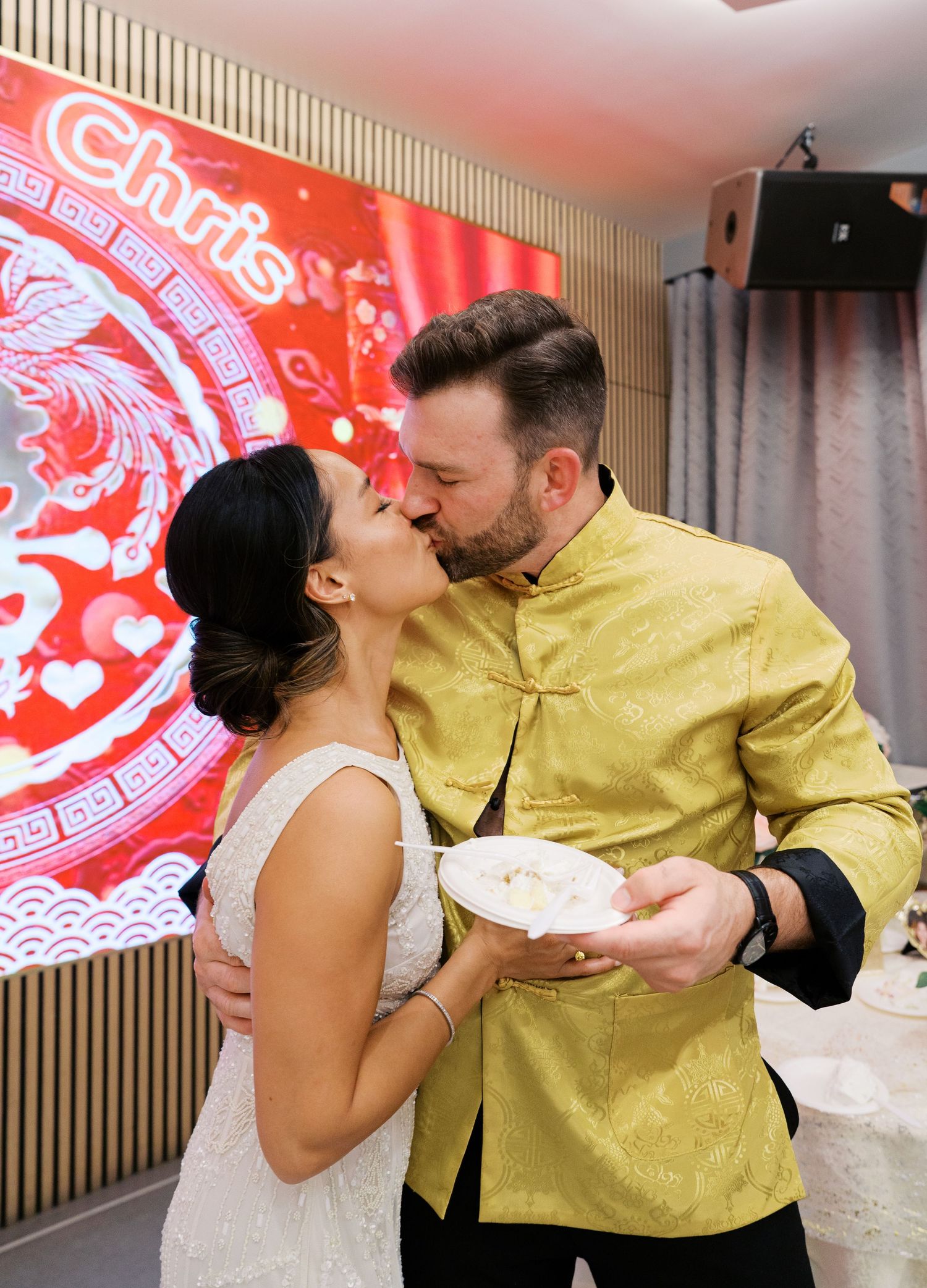 A couple shares a kiss while holding a plate in front of a red Chinese-themed backdrop.