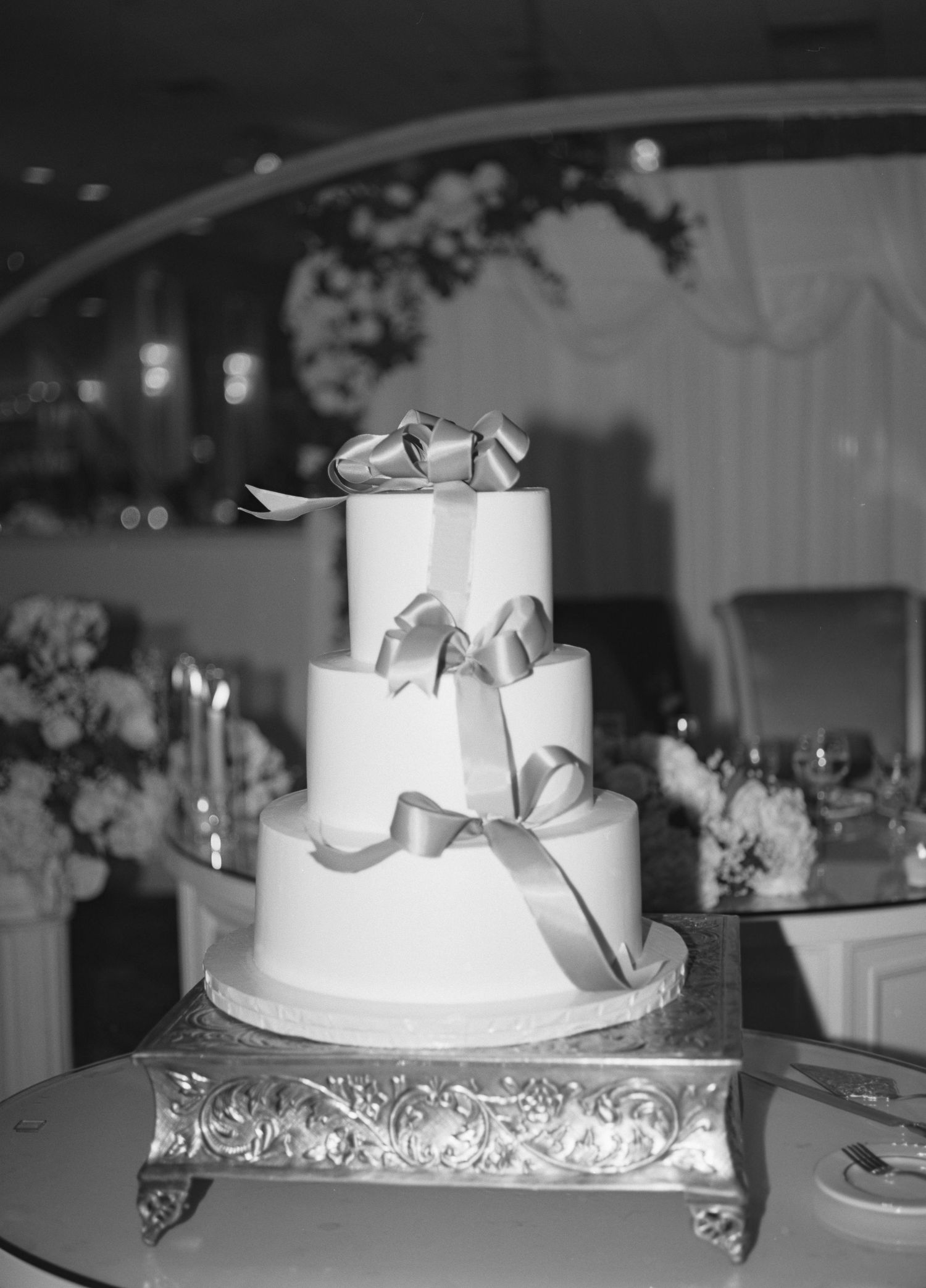 Elegant three-tiered white wedding cake with silver ribbon decorations displayed on an ornate metal stand.