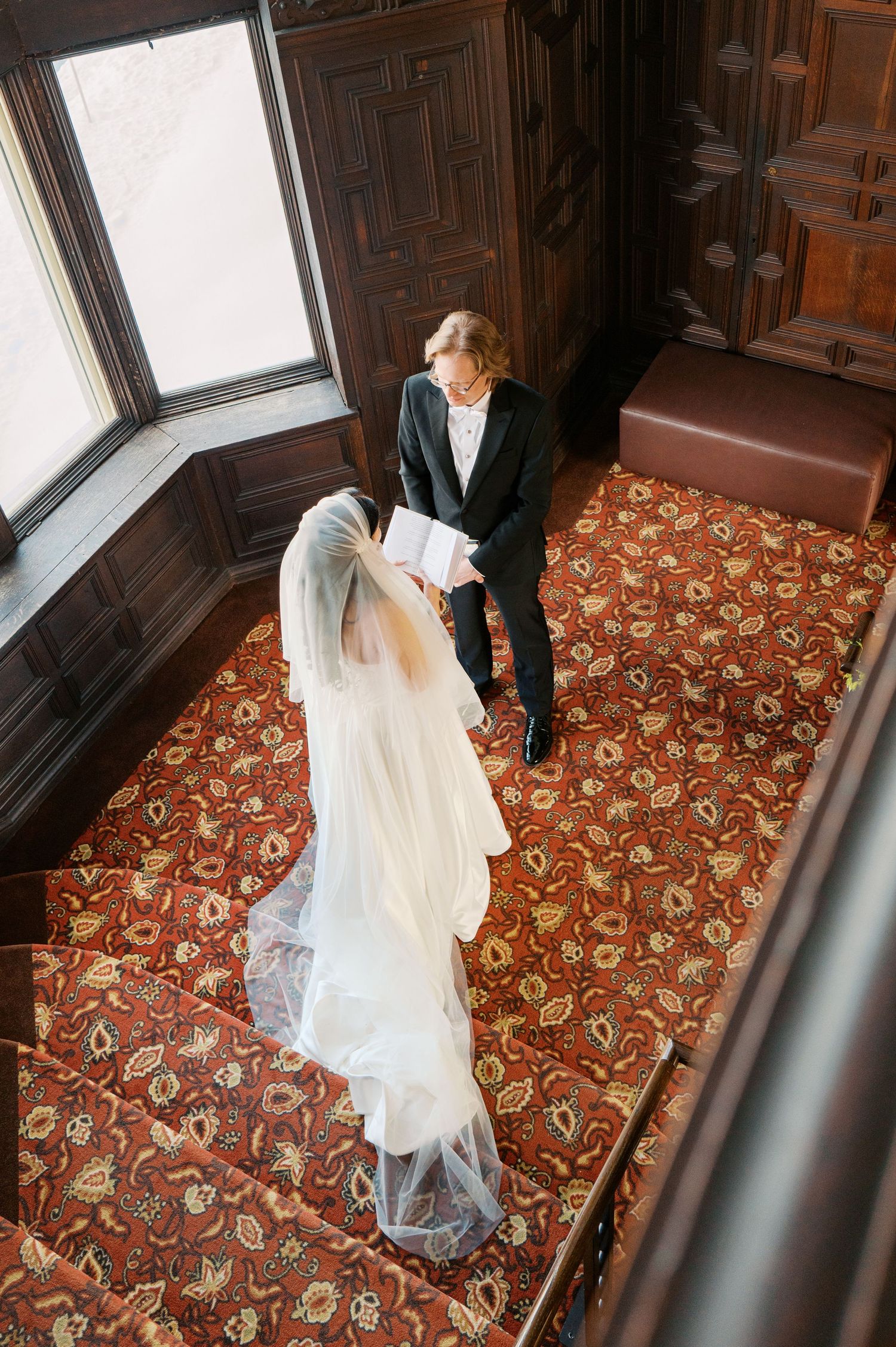 A couple in formal wedding attire share an intimate moment on ornate carpeted stairs near a bright window.