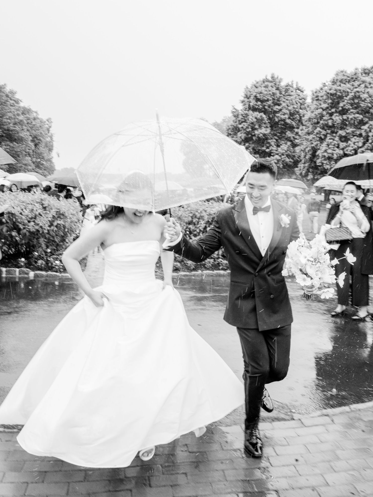 Black and white photo of a bride and groom sharing a clear umbrella, walking joyfully in the rain on their wedding day.