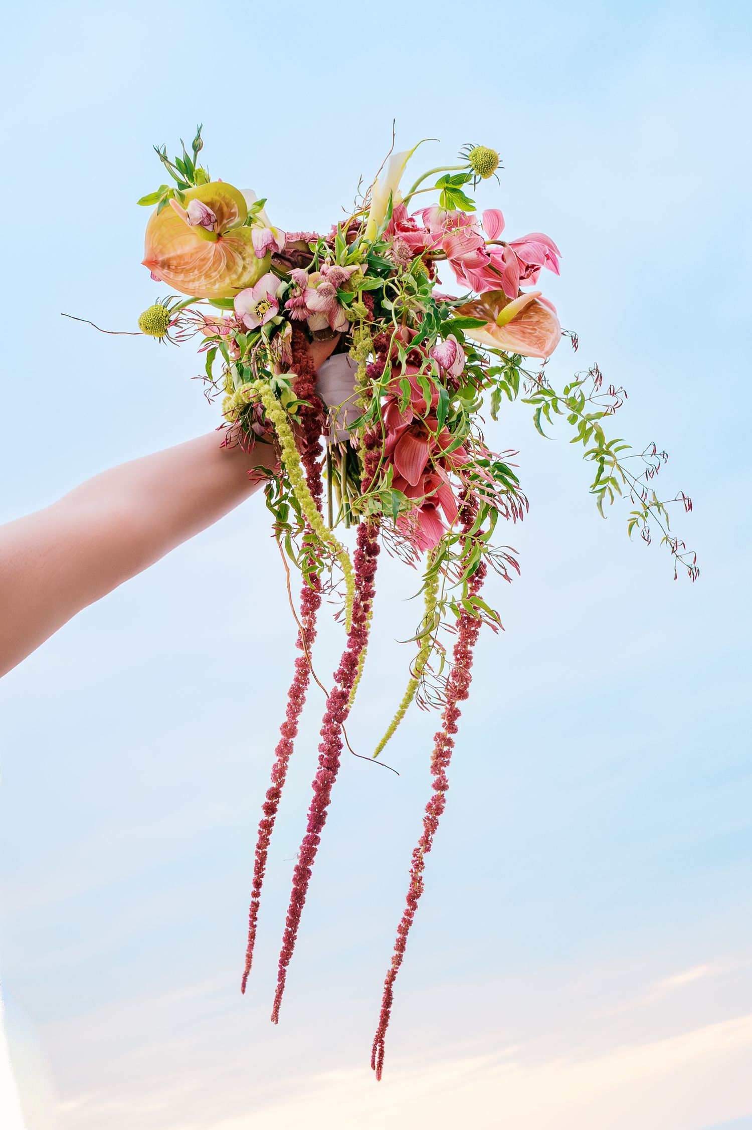 Delicate pink floral arrangement with cascading amaranth stems and greenery held against a soft sky background.