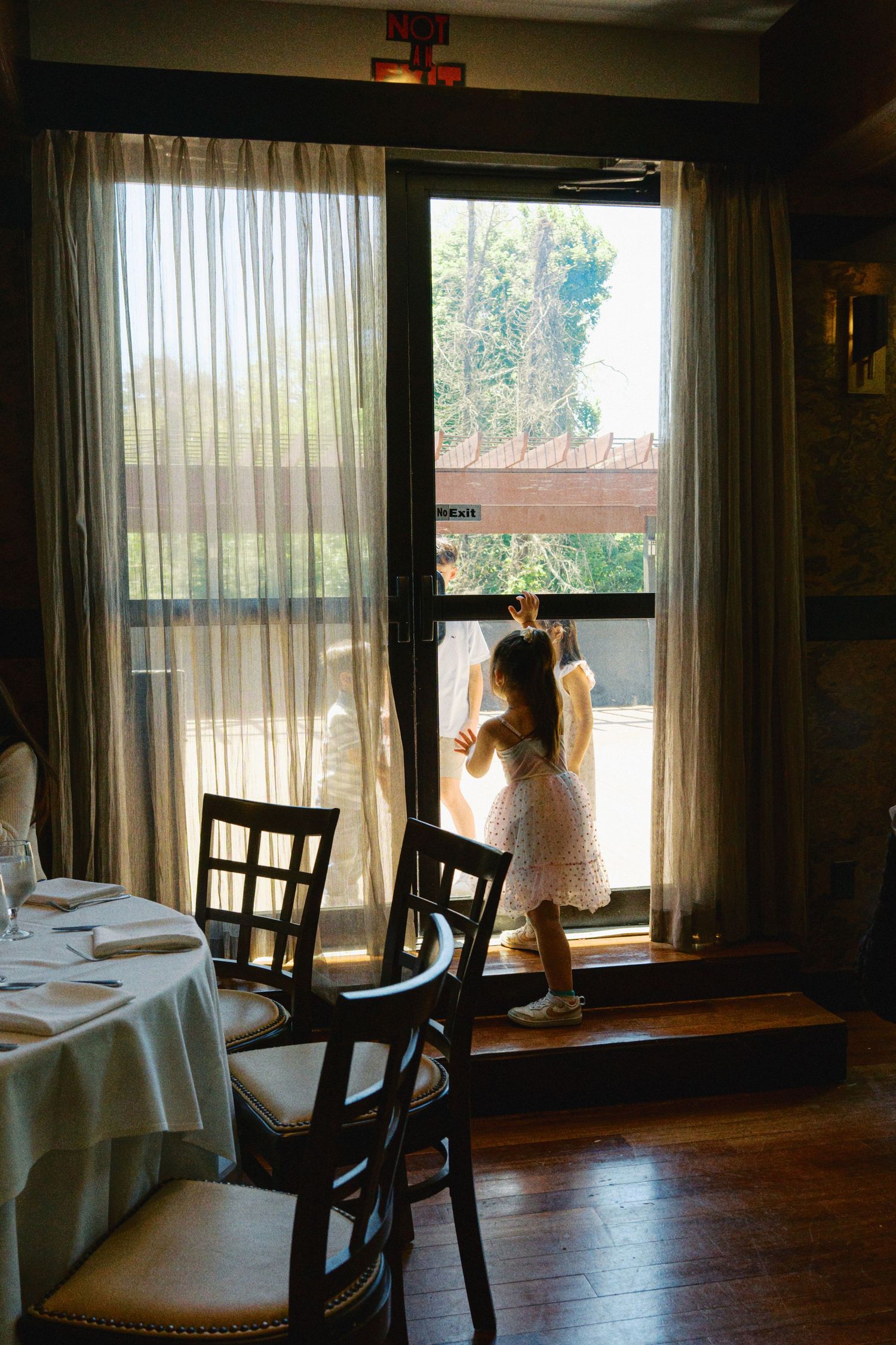 Young girl in white dress standing by large sunlit window in elegant restaurant dining room.