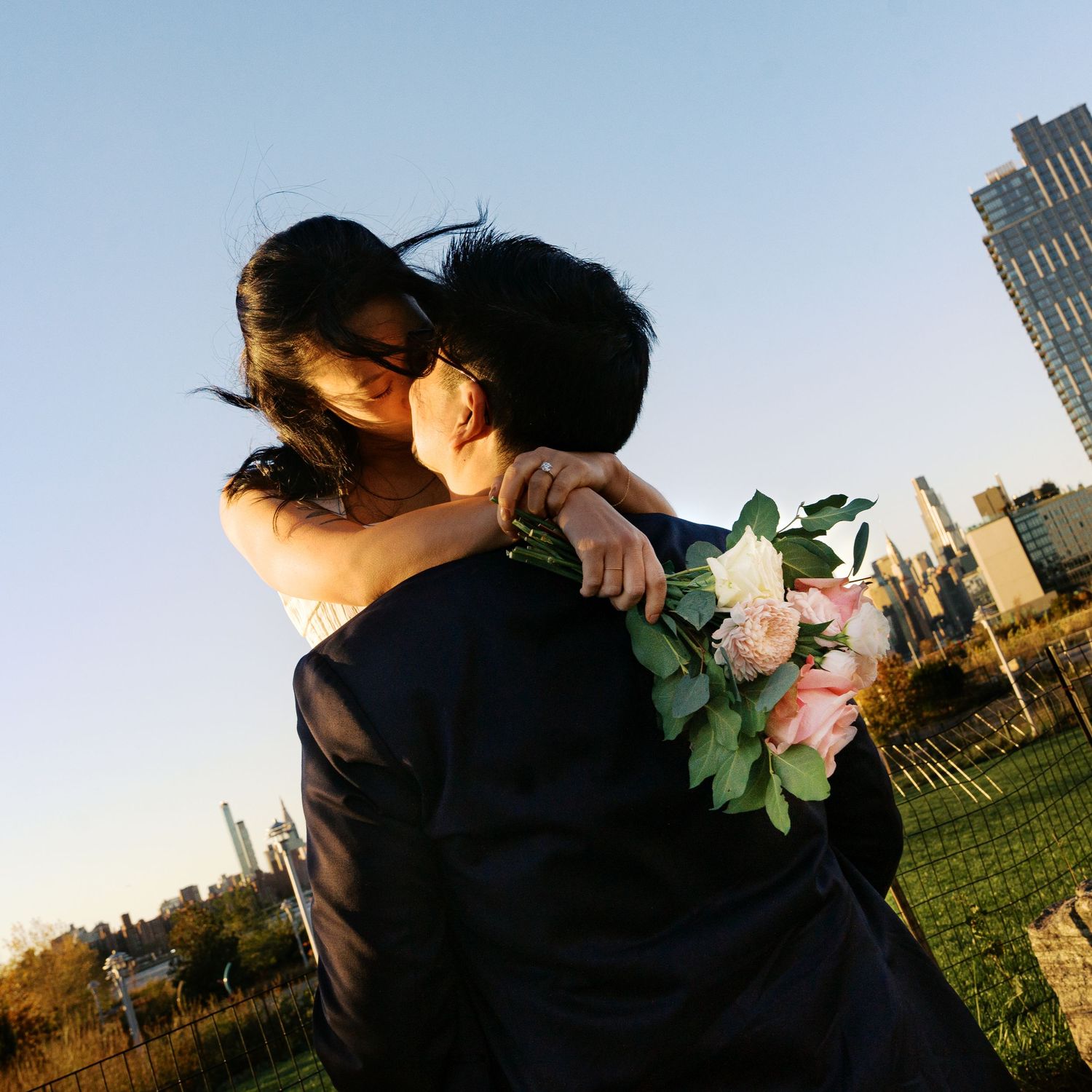 Couple embracing in a park with city skyscrapers visible in the background.