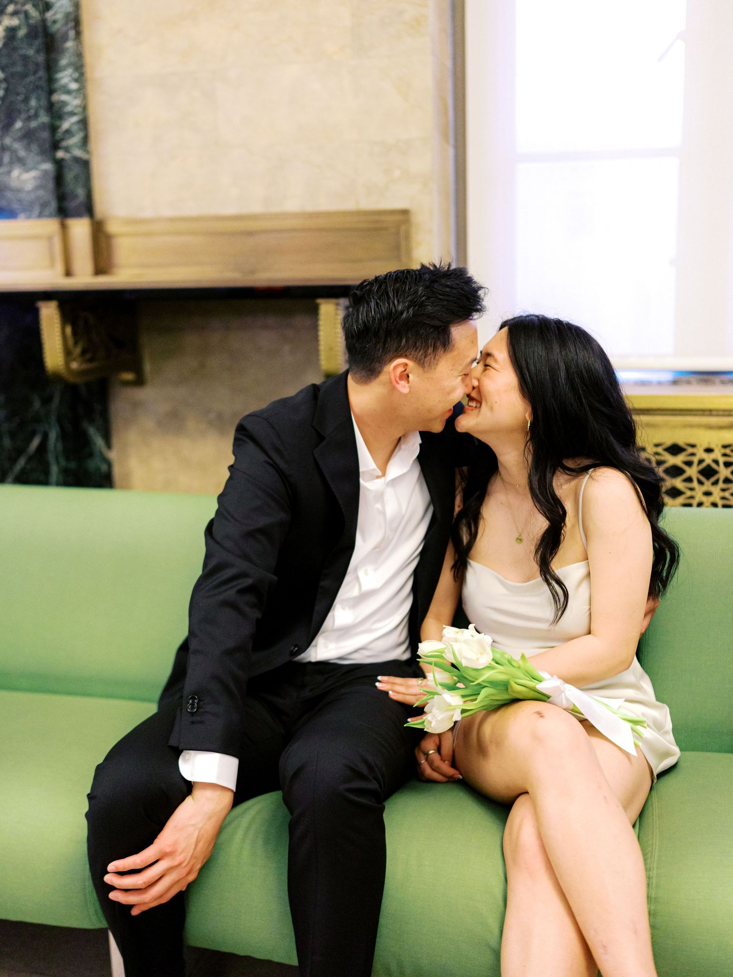 Couple sharing a tender kiss on a green sofa, bride holding white tulips in elegant attire.