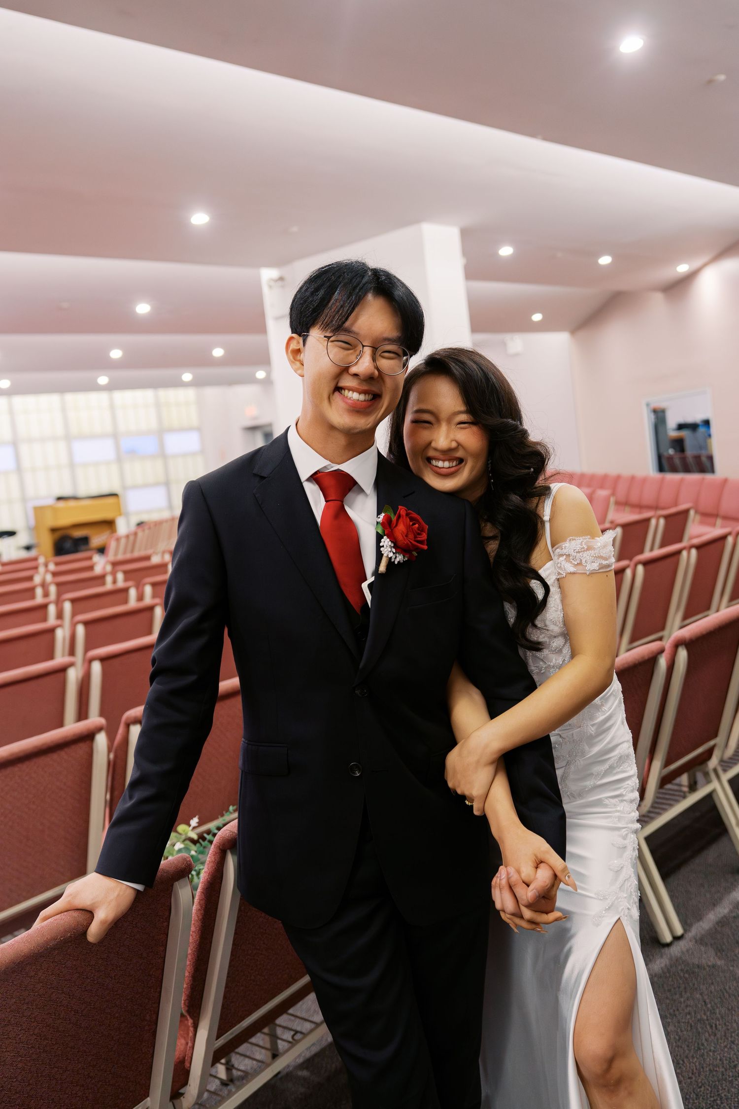 A couple poses playfully in a venue with rows of red chairs.