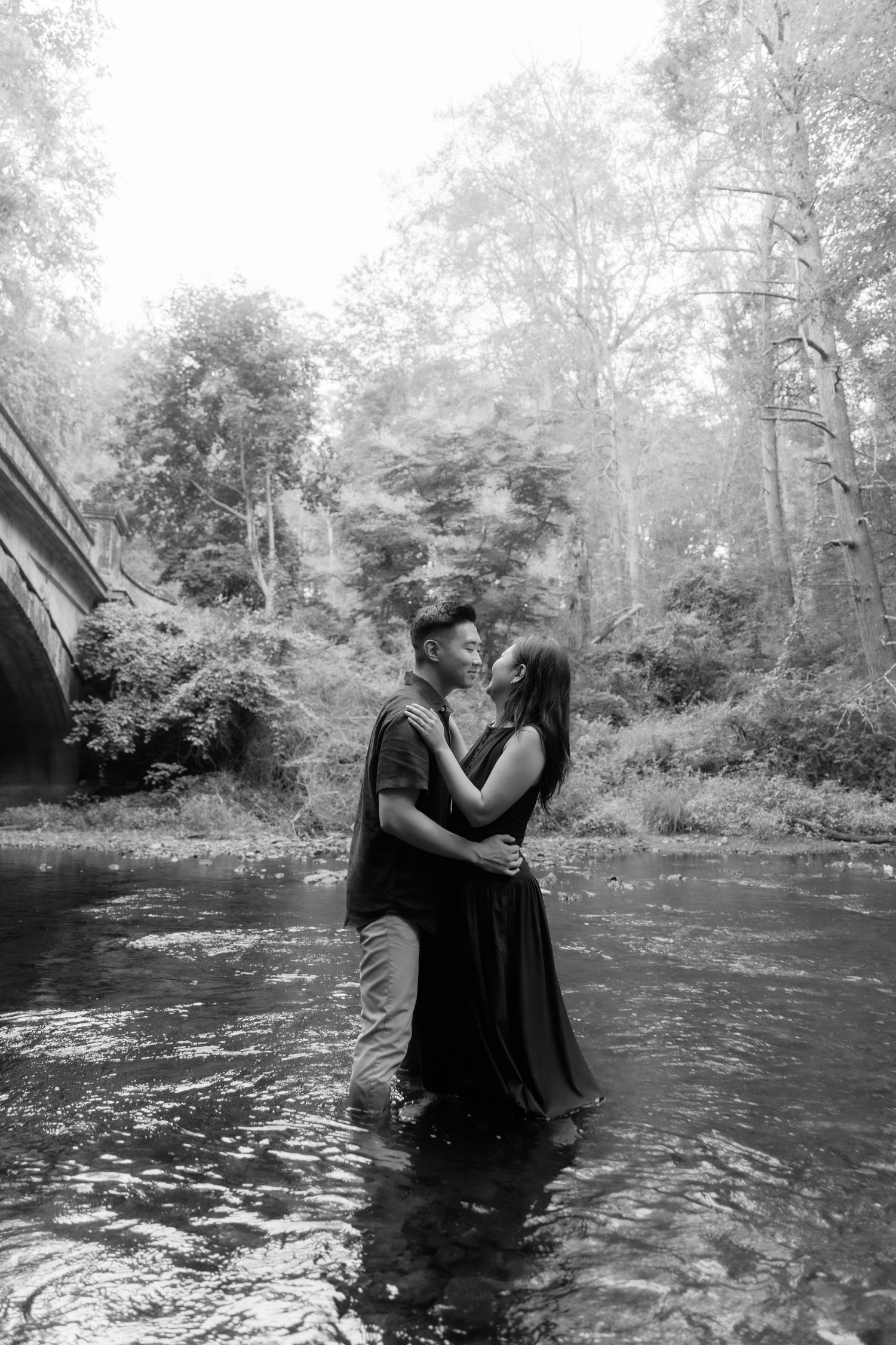 Two people embracing while standing in flowing water under trees.