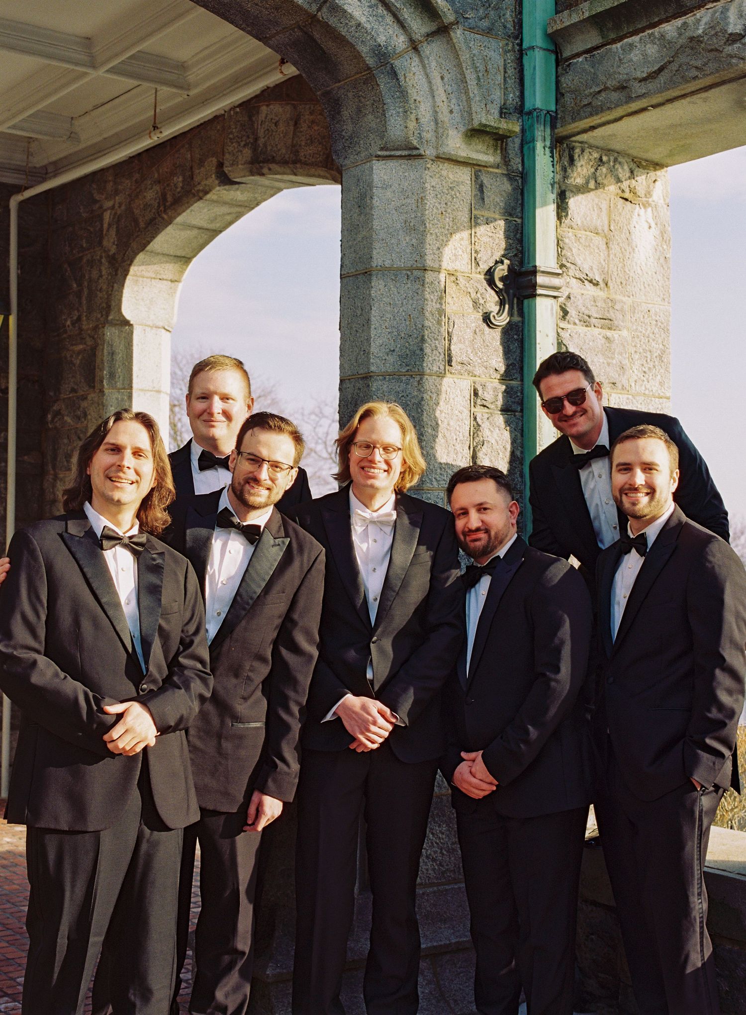 A group of groomsmen in black tuxedos pose together under a stone archway during a wedding celebration.