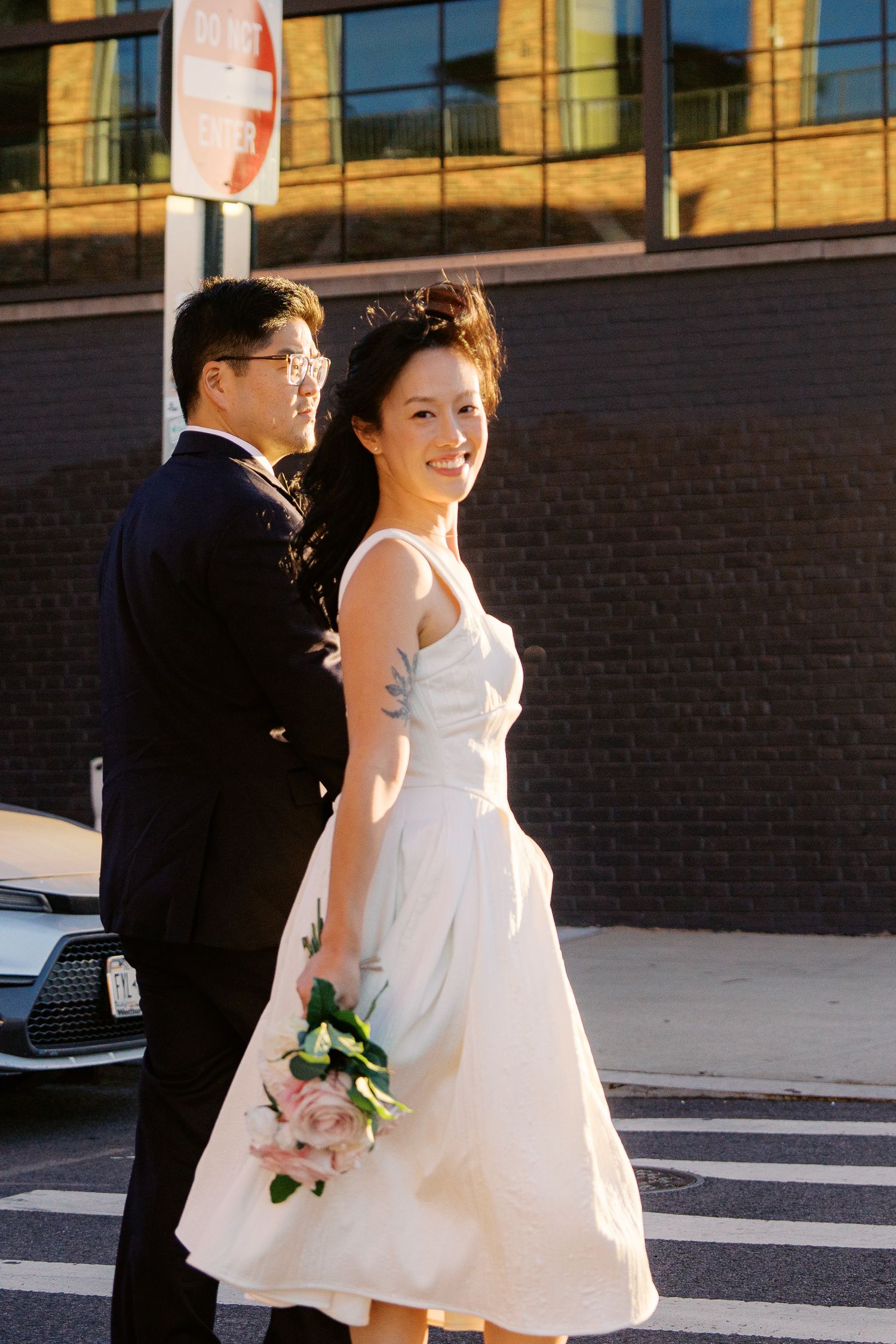 A flower girl in a white dress walks down a street while holding a small bouquet during a sunny day.