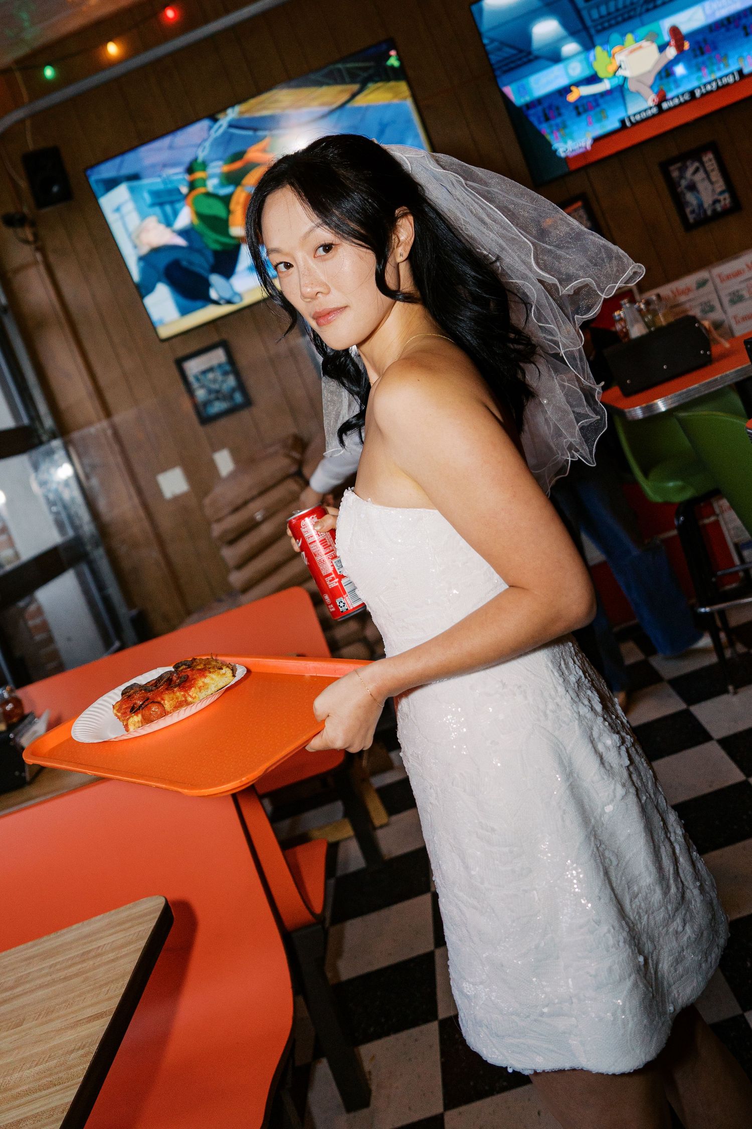 A bride in a white dress poses in a retro-style diner with orange booths.