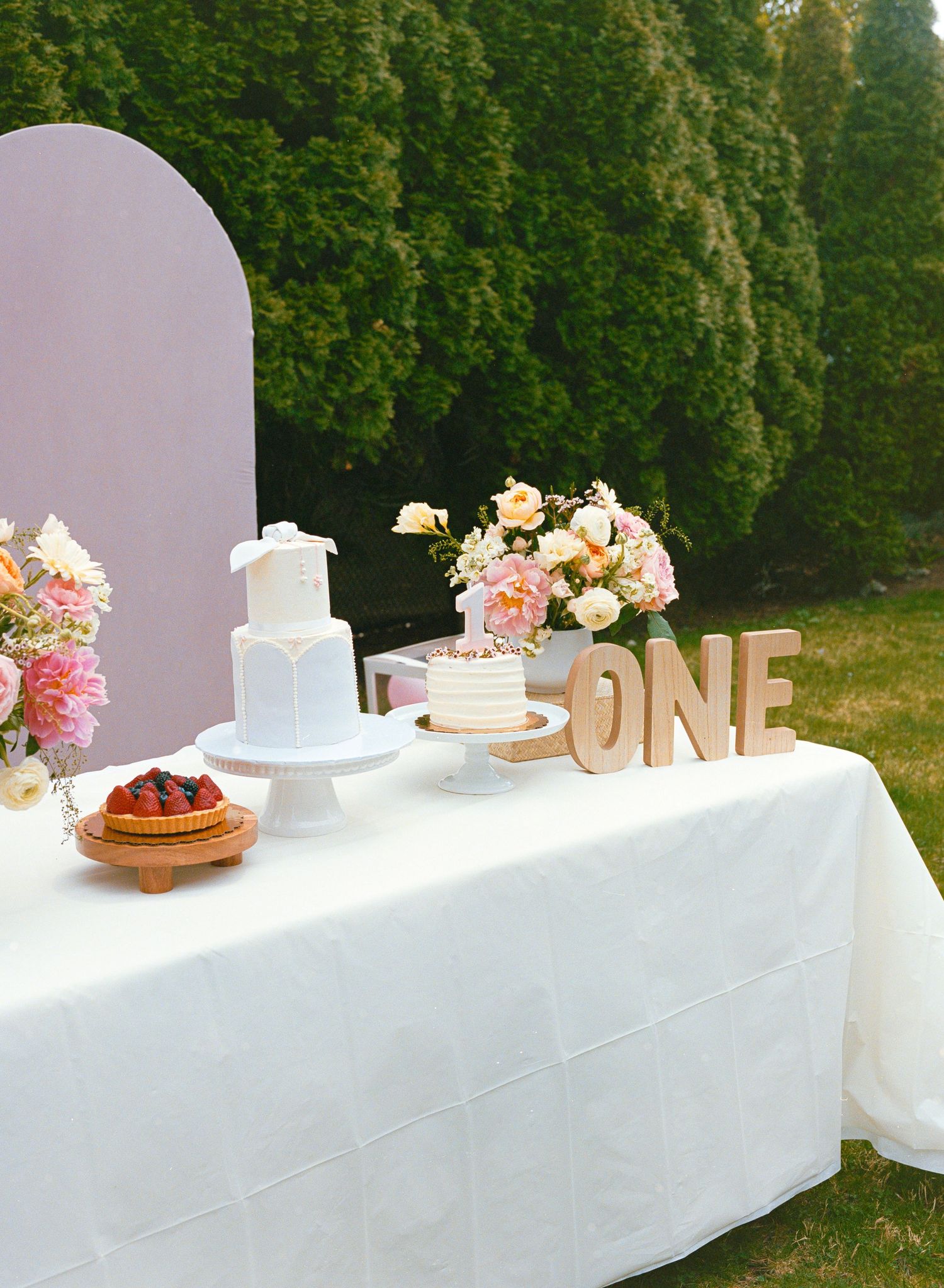 White dessert table display with cakes and decorative gold 'ONE' letters against a garden backdrop.