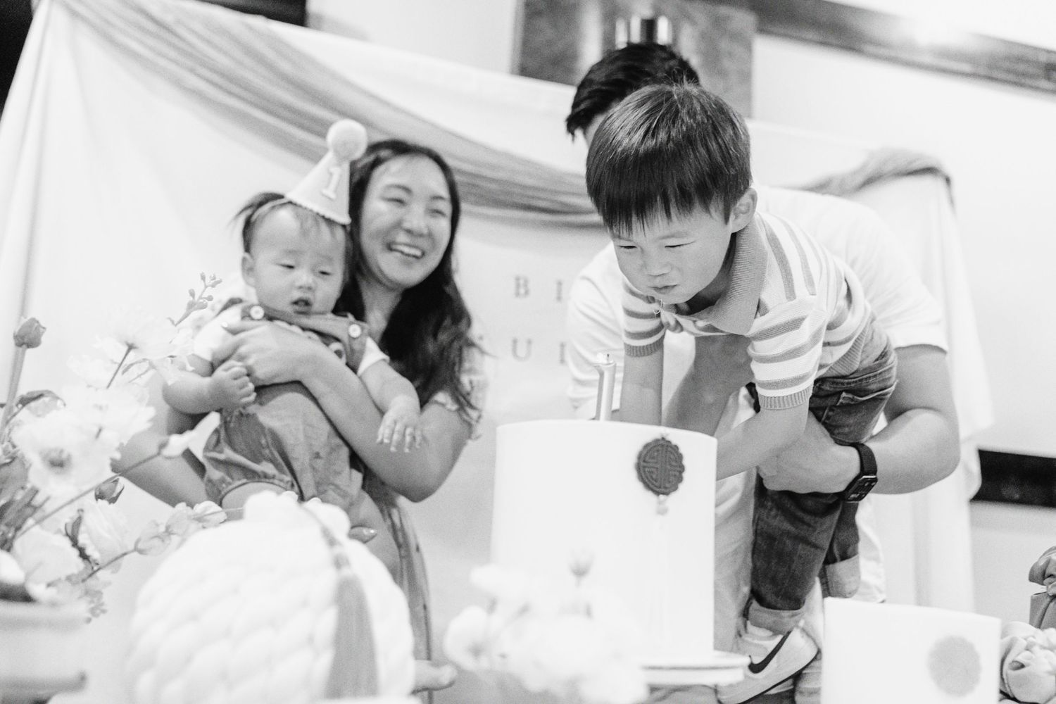 Black and white photo of a woman holding a baby and a child leaning over a birthday cake at a celebration.