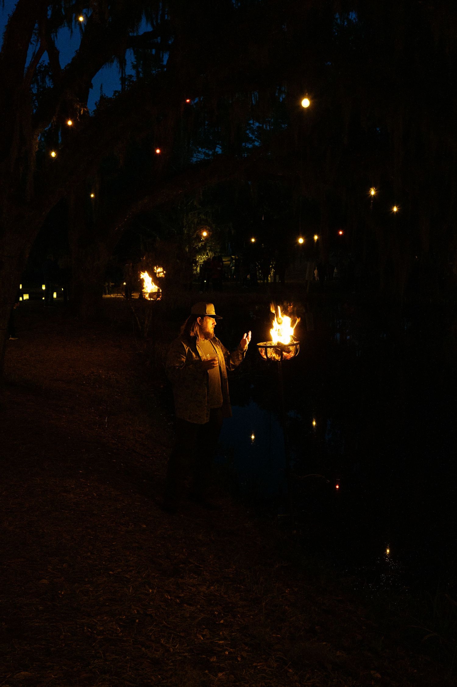 People gather around a campfire at night in a dark outdoor setting with trees visible in the background.
