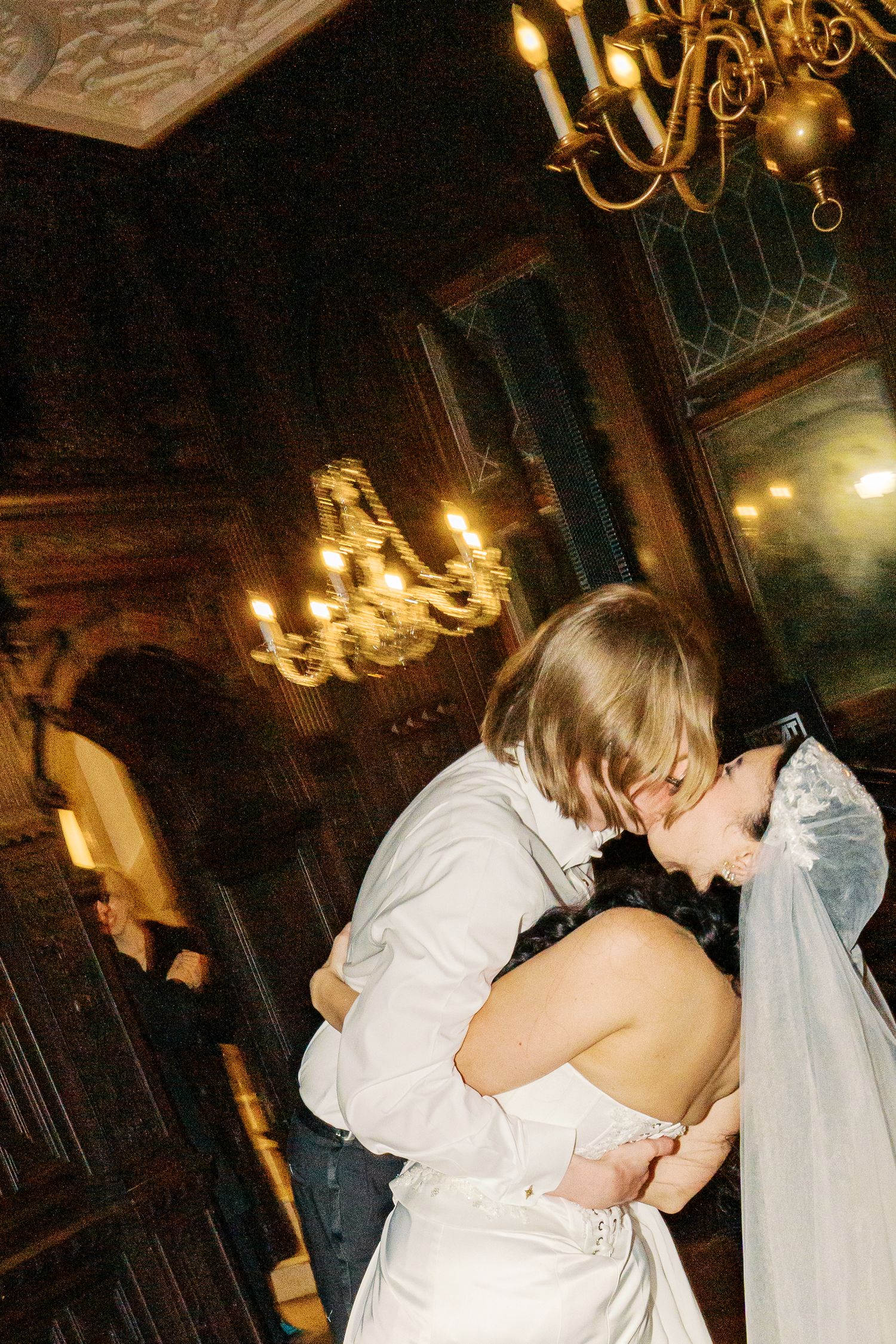 A couple shares their first dance in an elegant ballroom with ornate chandeliers and warm lighting.