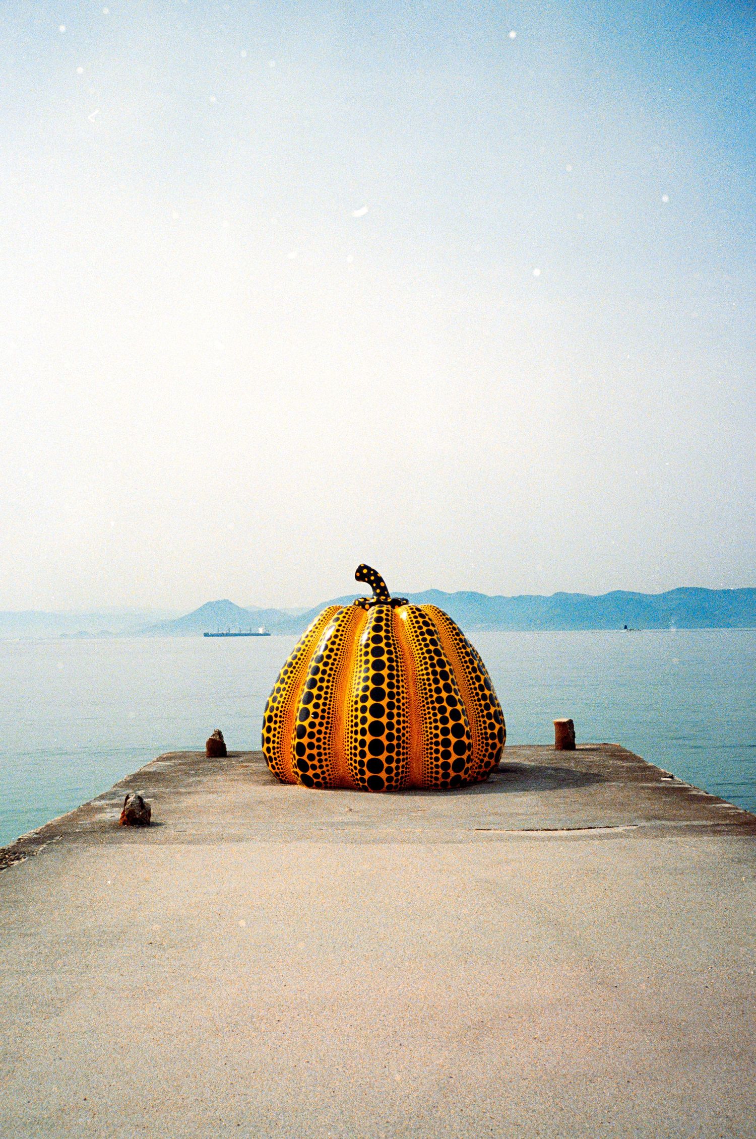 A large yellow and black polka dot pumpkin sculpture sits on a concrete pier overlooking the ocean.