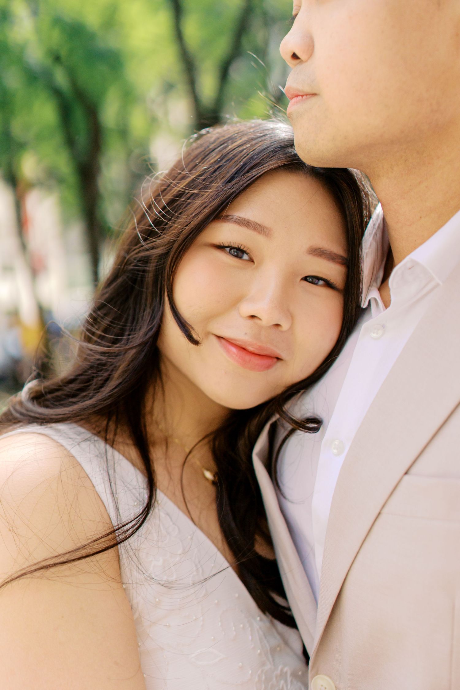 A young Asian woman smiles warmly, leaning against a man in a beige suit outdoors, in a romantic couple portrait.