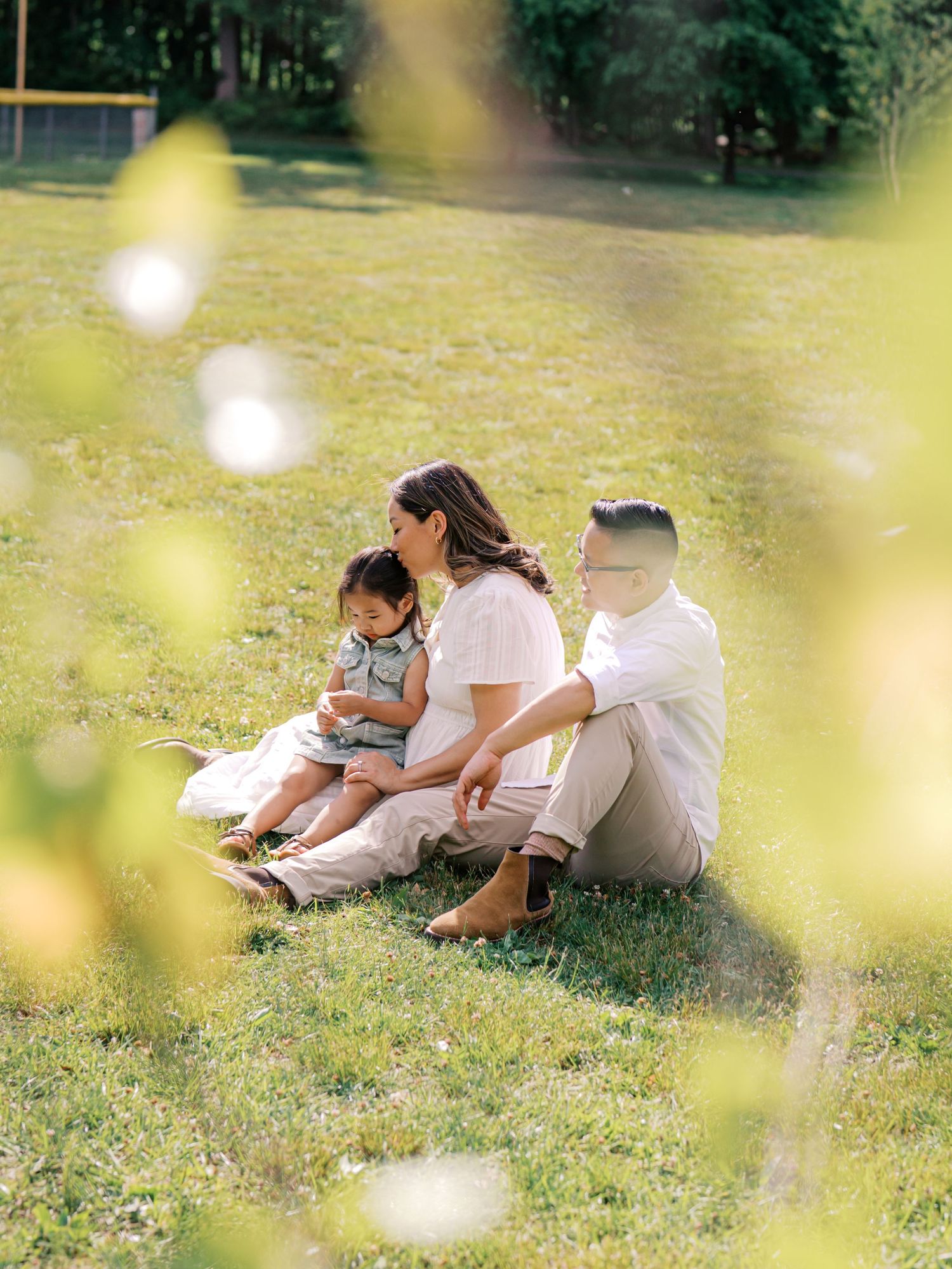 Family of three sitting on green grass in sunlit park, parents gazing at toddler in soft natural light.